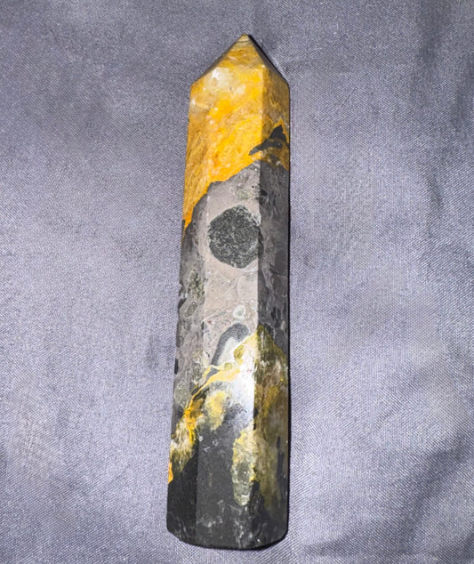 A natural pointed crystal tower, likely a jasper or quartz point, featuring a marbled pattern of bright yellow, dark gray, and black, resting on a textured gray fabric background.