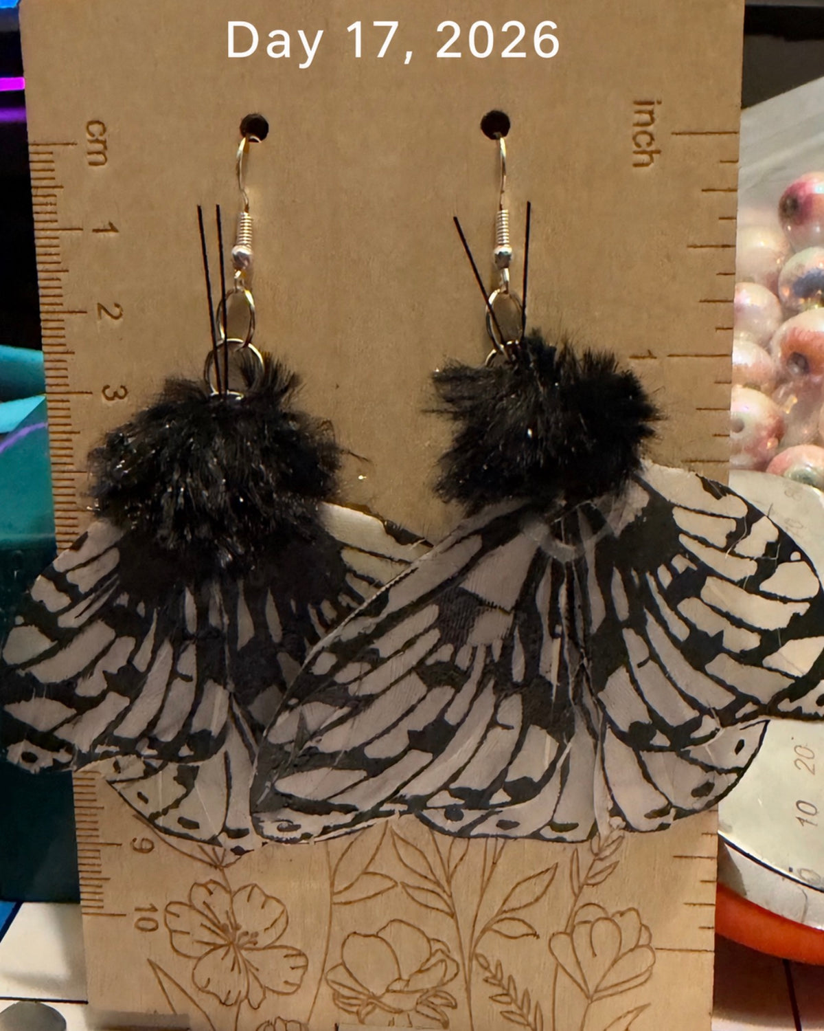 A pair of dangling earrings displayed on a cardboard stand with a ruler for scale. Each earring has a black-and-white patterned moth wing design with fluffy black pom-poms and silver fishhook ear wires.