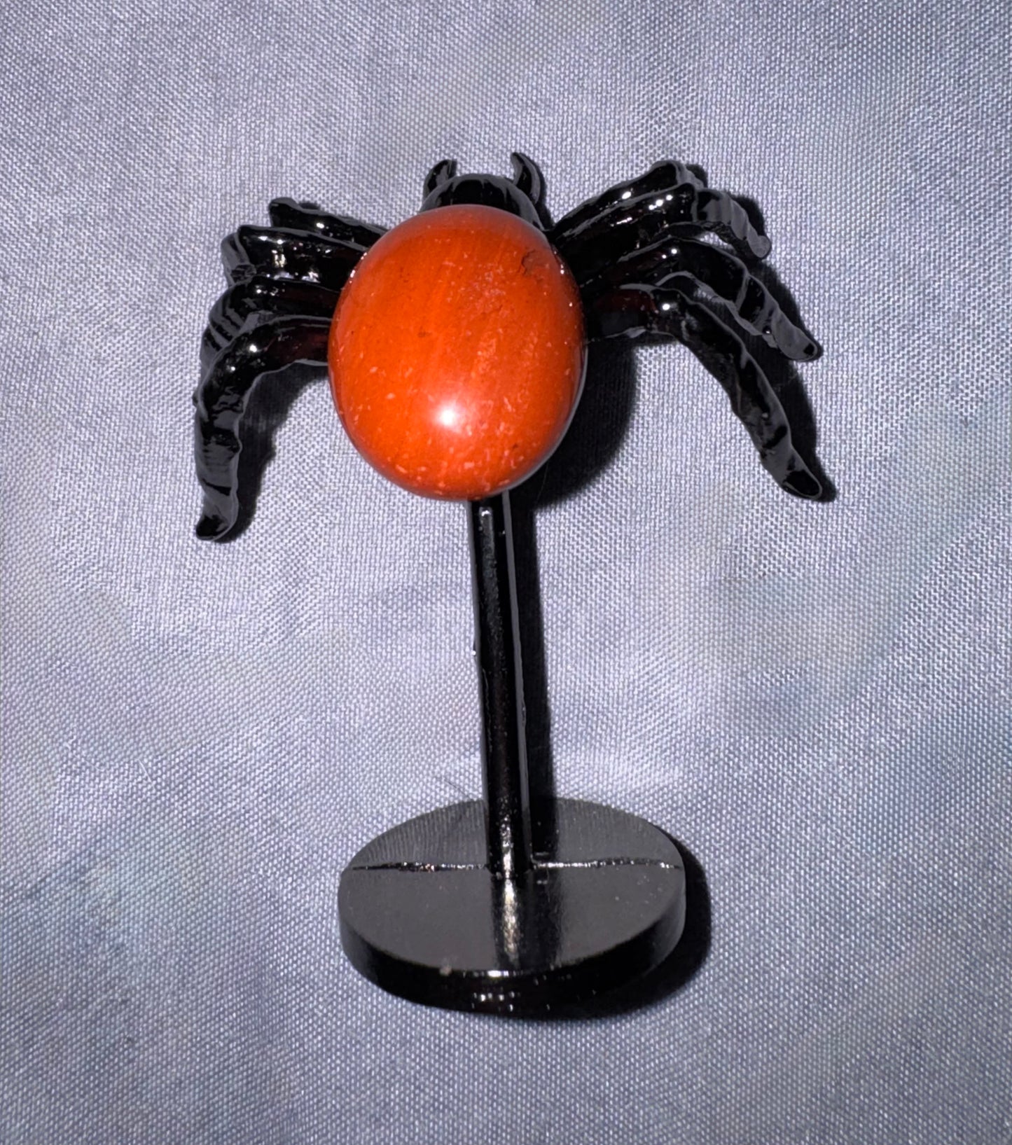 A small black spider-shaped decorative object with a round orange-red center, displayed on a black stand against a light gray fabric background.