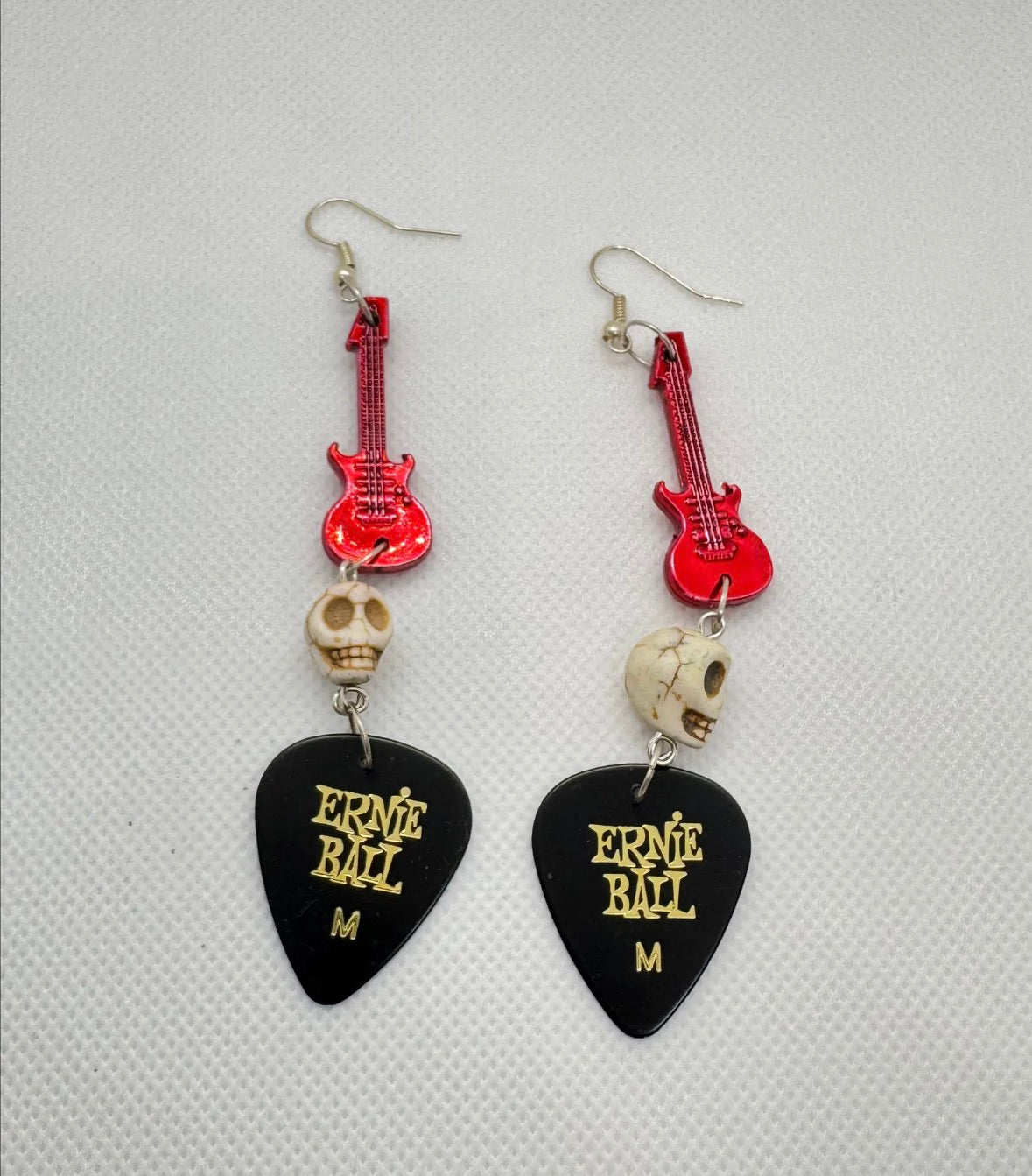 Guitar pick earrings with red guitar-shaped charms and skull beads on a gray background