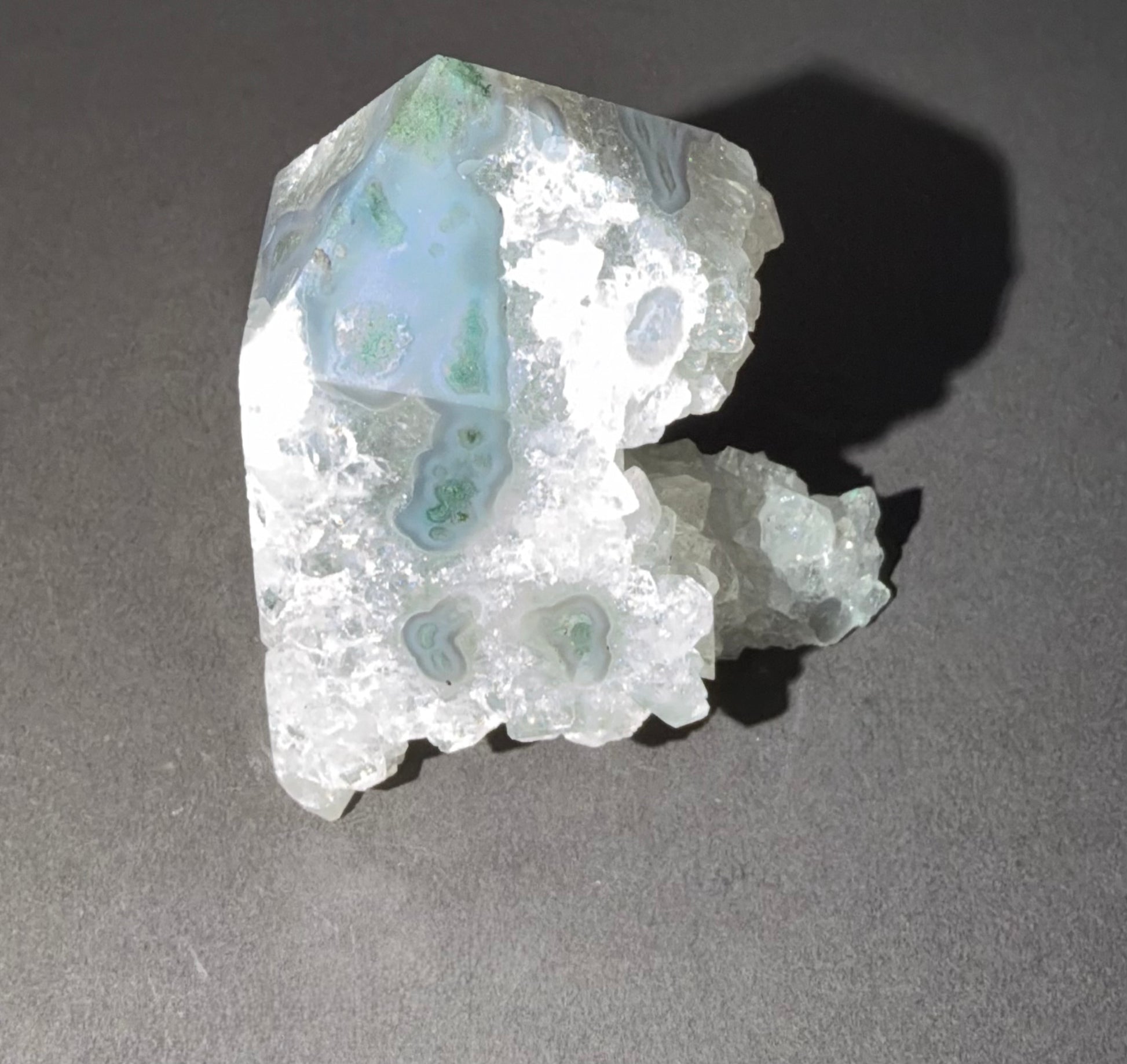 A raw agate crystal specimen with light blue and green banding patterns, set against a dark gray background with a sharp shadow.