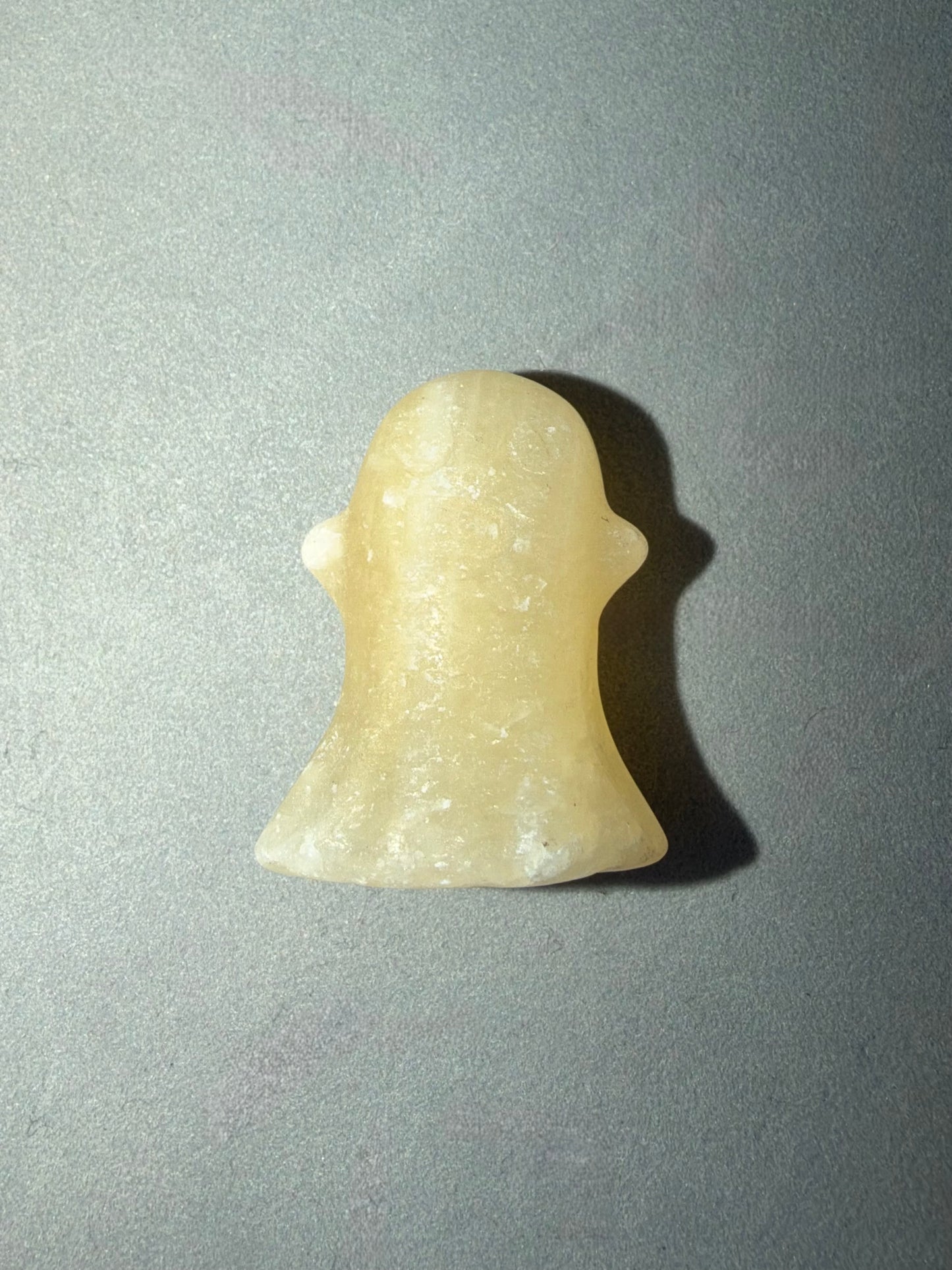 A small ghost-shaped pendant or charm made of pale translucent stone, photographed from the back on a gray background with a cast shadow.