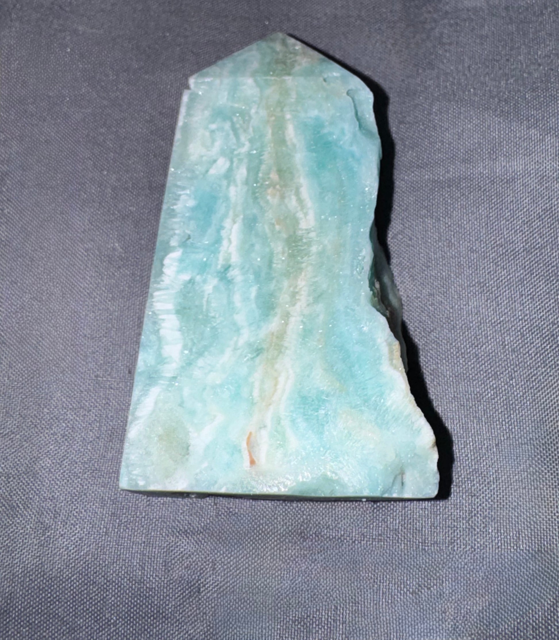A polished natural stone chisel with a pointed top, featuring light blue-green and white marbled patterns, resting on a textured gray fabric background.