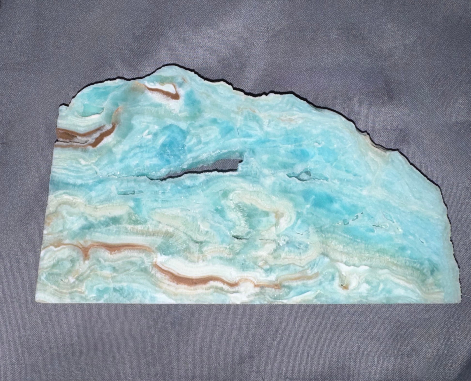 A raw, polished slab of turquoise-colored stone with natural marbled patterns and brown veining, displayed on a textured gray background.