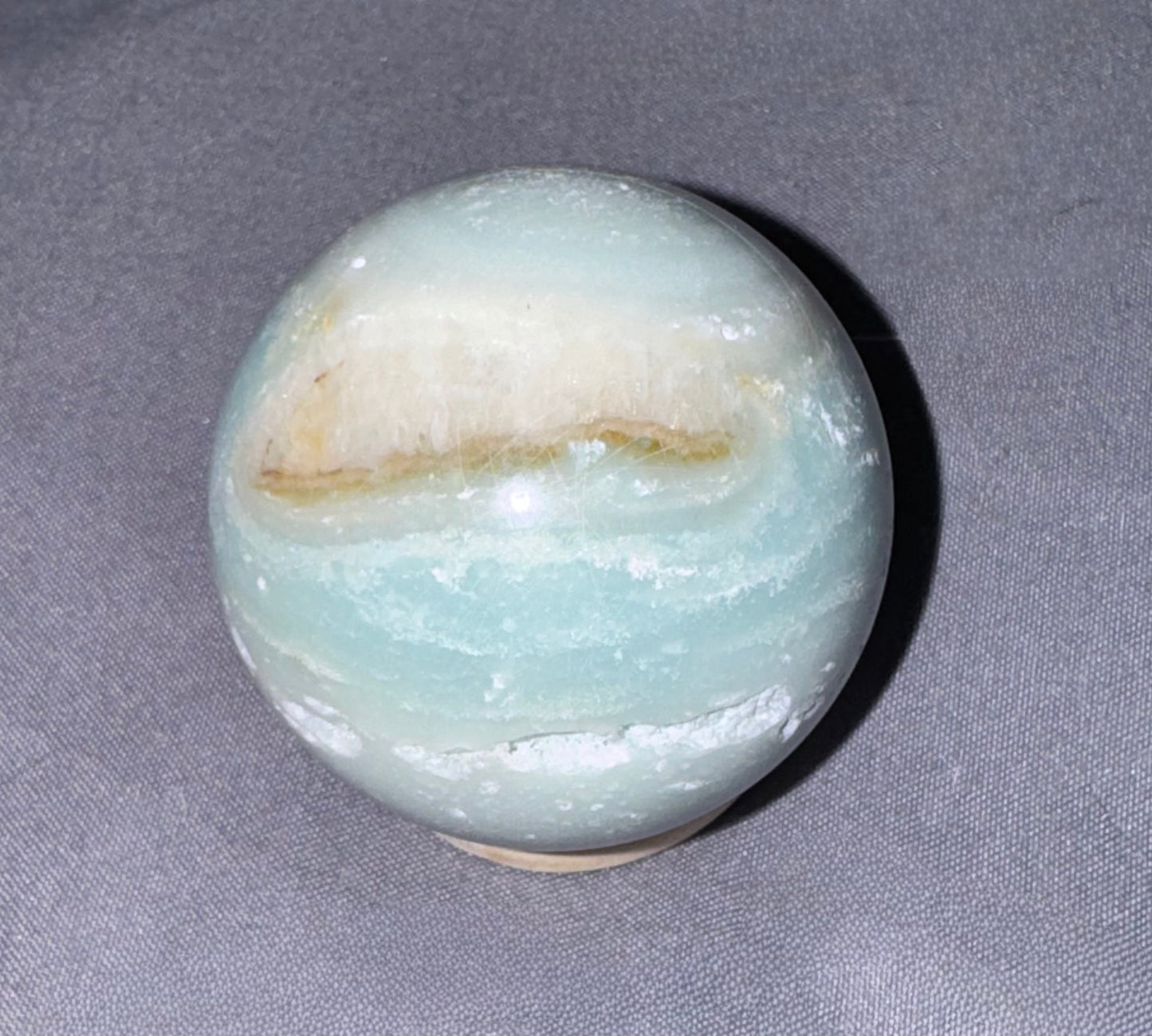 A polished round stone sphere, likely amazonite, featuring pale green-blue bands with a cream and light brown band near the top, resting on a gray textured fabric background.