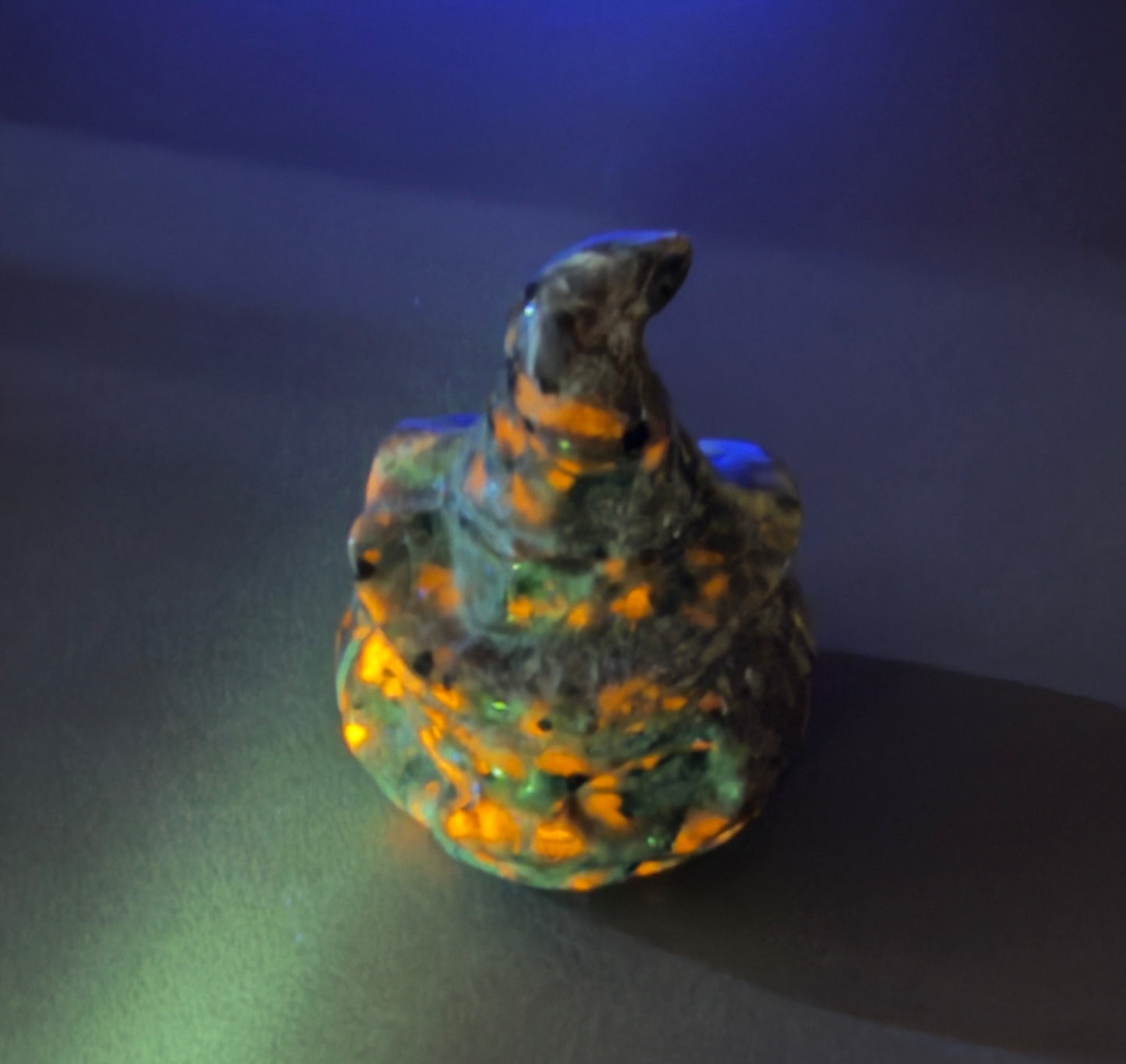 A small sculpted figurine resembling a bird or creature with a pointed crest, shown under UV lighting that makes it glow with orange, green, and blue colors against a dark background.