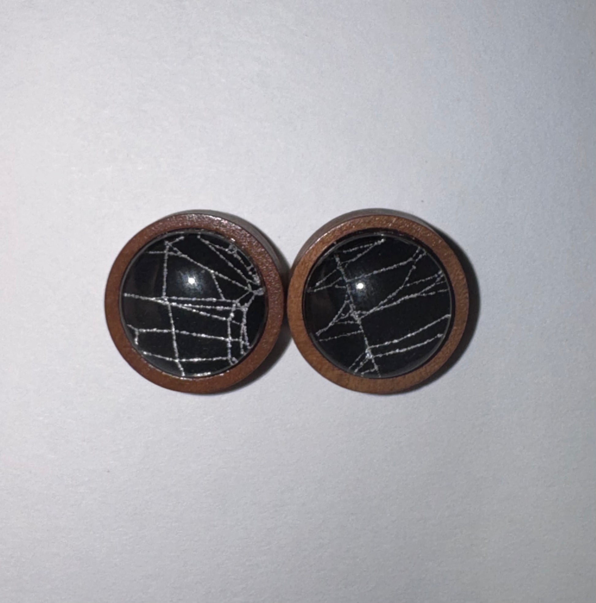 A pair of round wooden button covers with black centers featuring a white geometric line pattern, set against a plain light gray background.