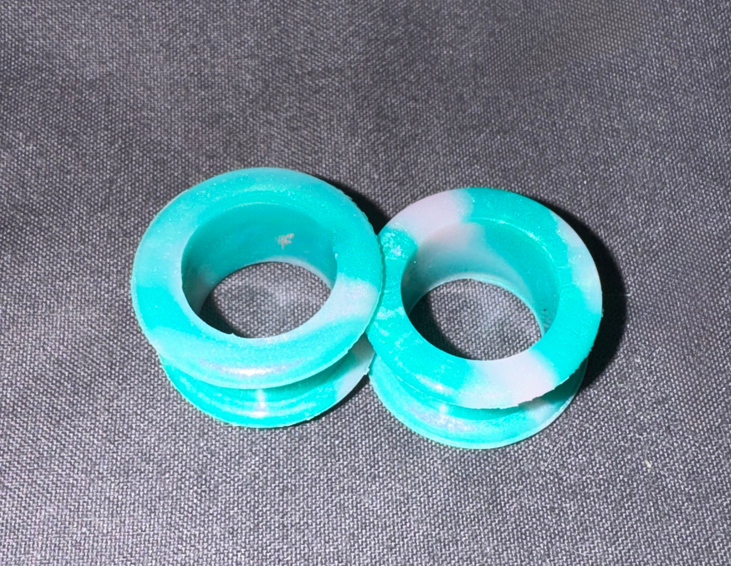 Two turquoise and white marbled acrylic ear plugs on a dark gray textured background.