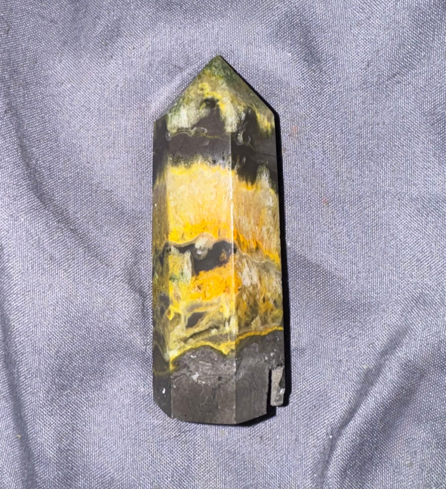 A natural pointed crystal tower, likely a quartz point, featuring a marbled pattern of yellow, black, and gray, resting on a textured gray fabric background.