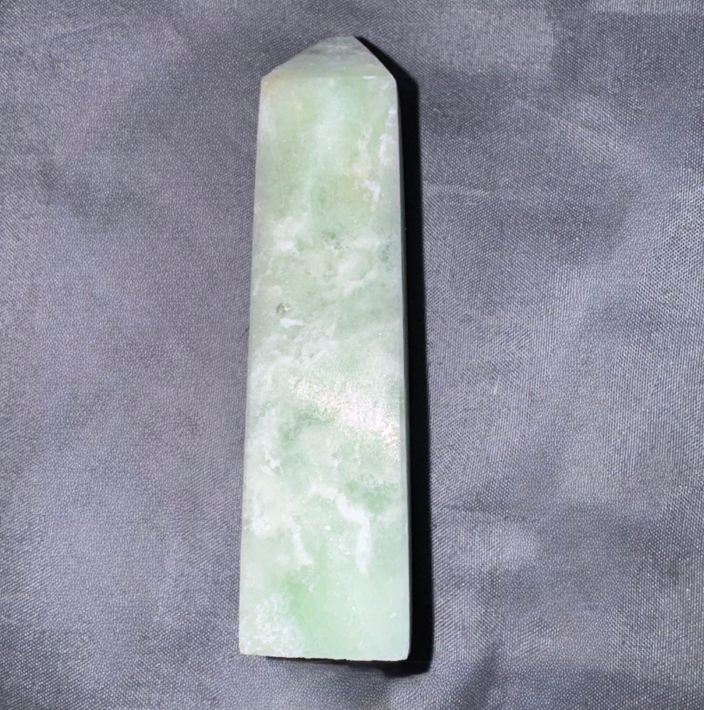 A natural green jadeite crystal point wand with a smooth polished surface, standing vertically on a textured gray fabric background.