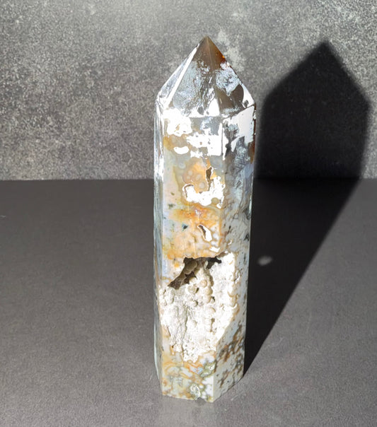 A polished natural crystal point, featuring a mix of white, gray, and orange-brown mineral inclusions, standing on a dark gray surface with a textured gray background.
