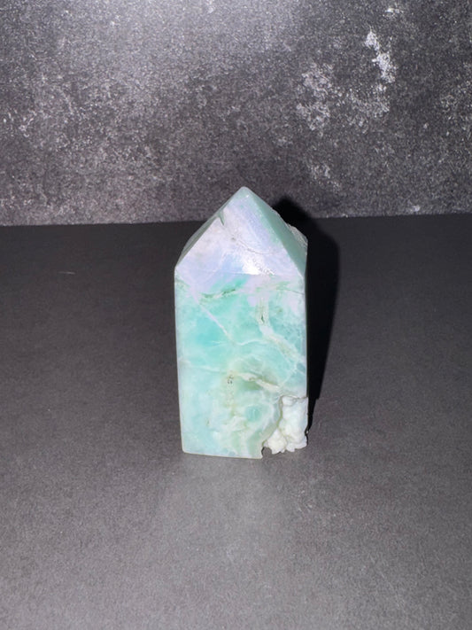 A natural  crystal tower with a pointed top, showing light green and white marbled patterns, set against a dark gray background.