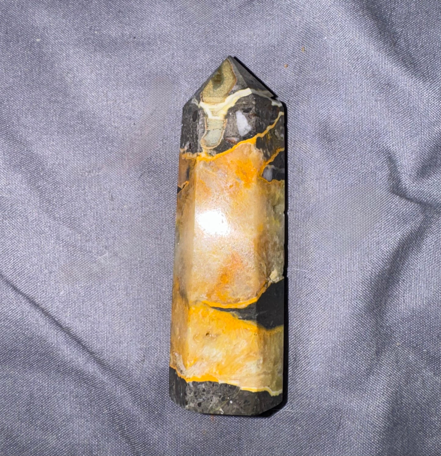 A natural crystal point with a marbled pattern of black, orange, and cream, photographed on a textured gray fabric background.