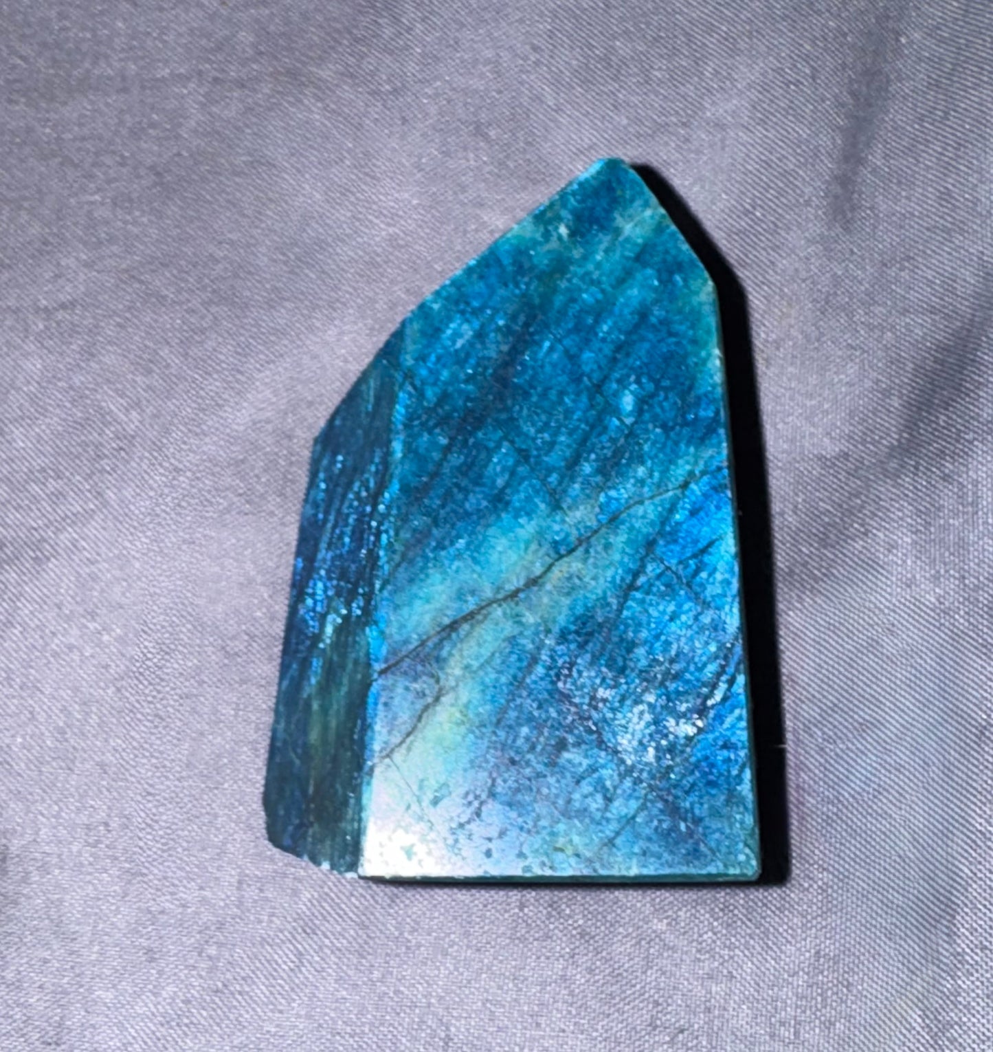 A polished blue labradorite crystal point with natural natural banding, resting on a textured gray fabric surface.