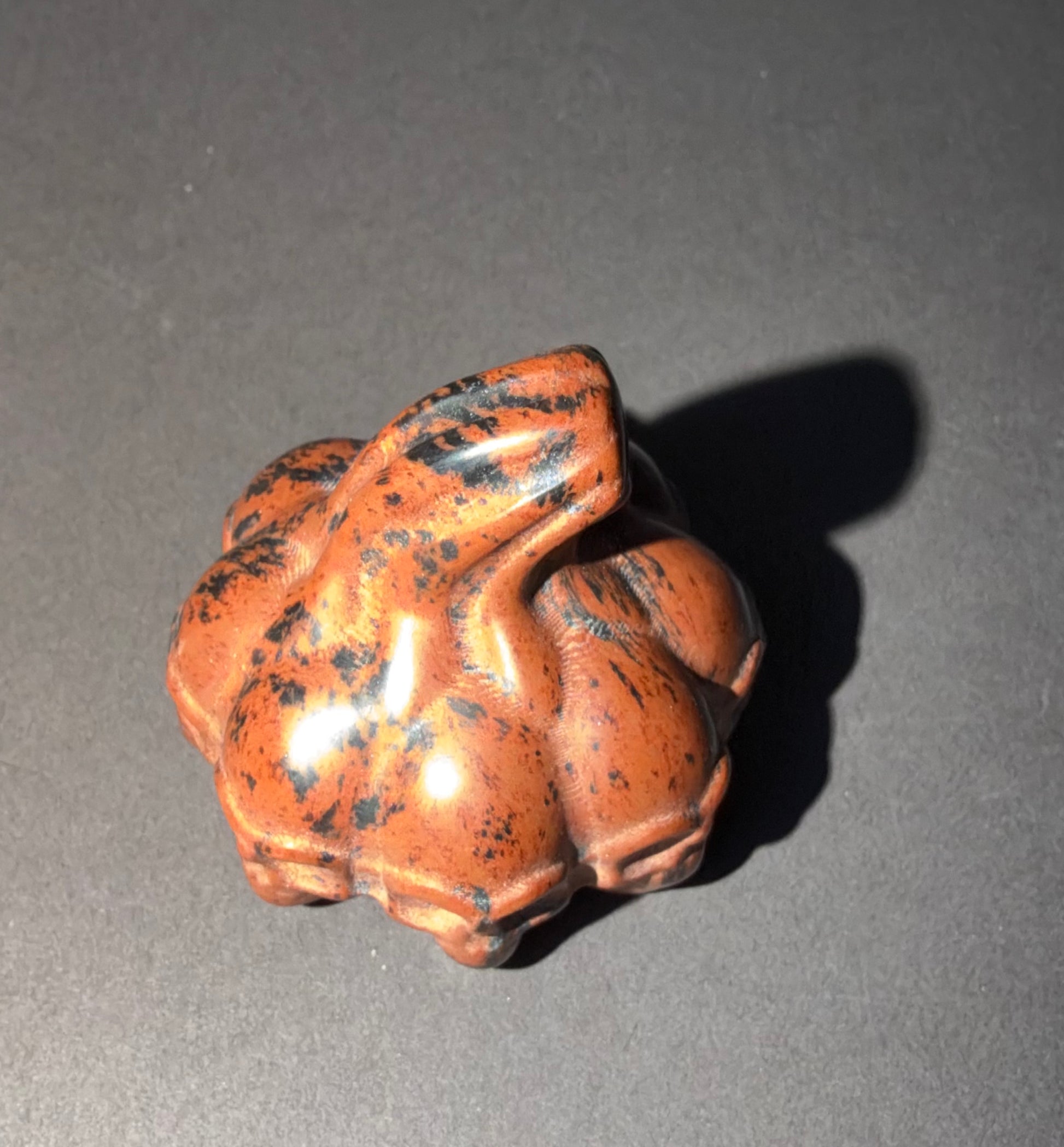 A small carved figurine of a frog in a reddish-brown stone with black speckles, photographed on a dark gray surface with a cast shadow.