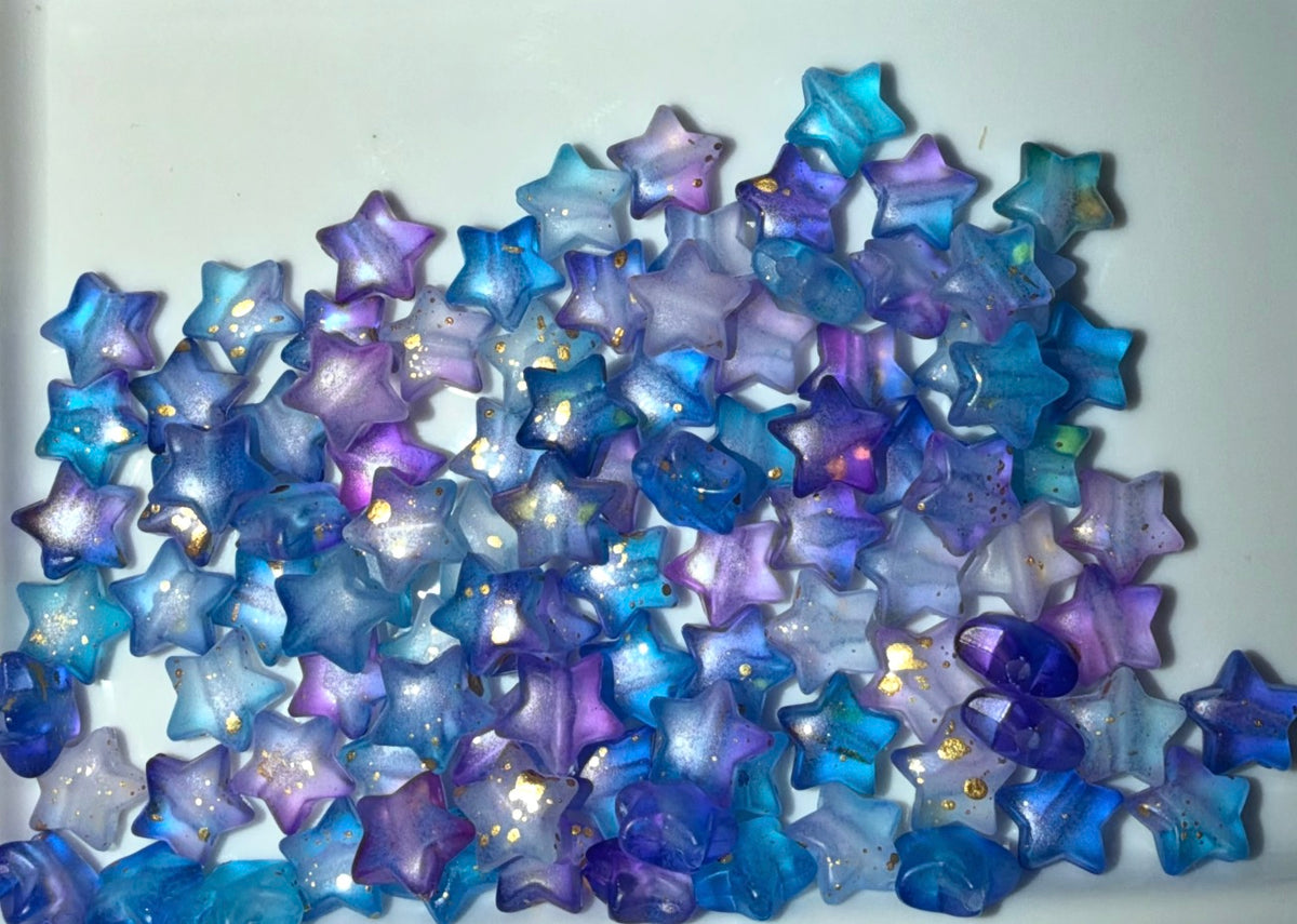 A pile of small translucent star-shaped beads in shades of blue, purple, and lavender, some with tiny gold foil flecks, on a plain white background.