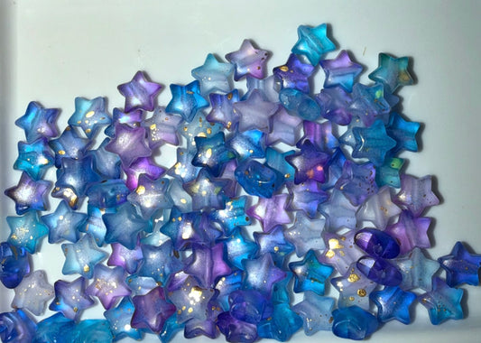 A pile of small translucent star-shaped beads in shades of blue, purple, and lavender, some with tiny gold foil flecks, on a plain white background.