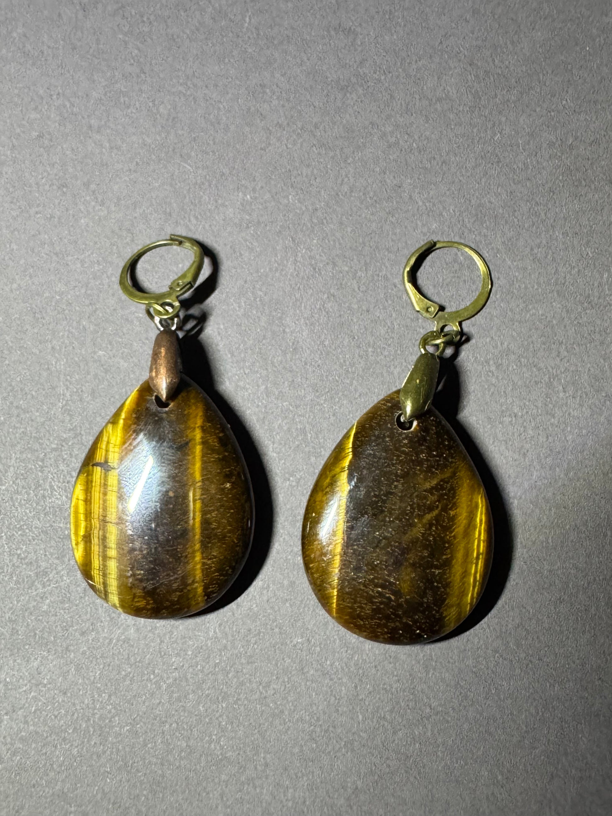 A pair of teardrop-shaped tiger's eye drop earrings with gold leverback hooks, displayed on a neutral gray background.