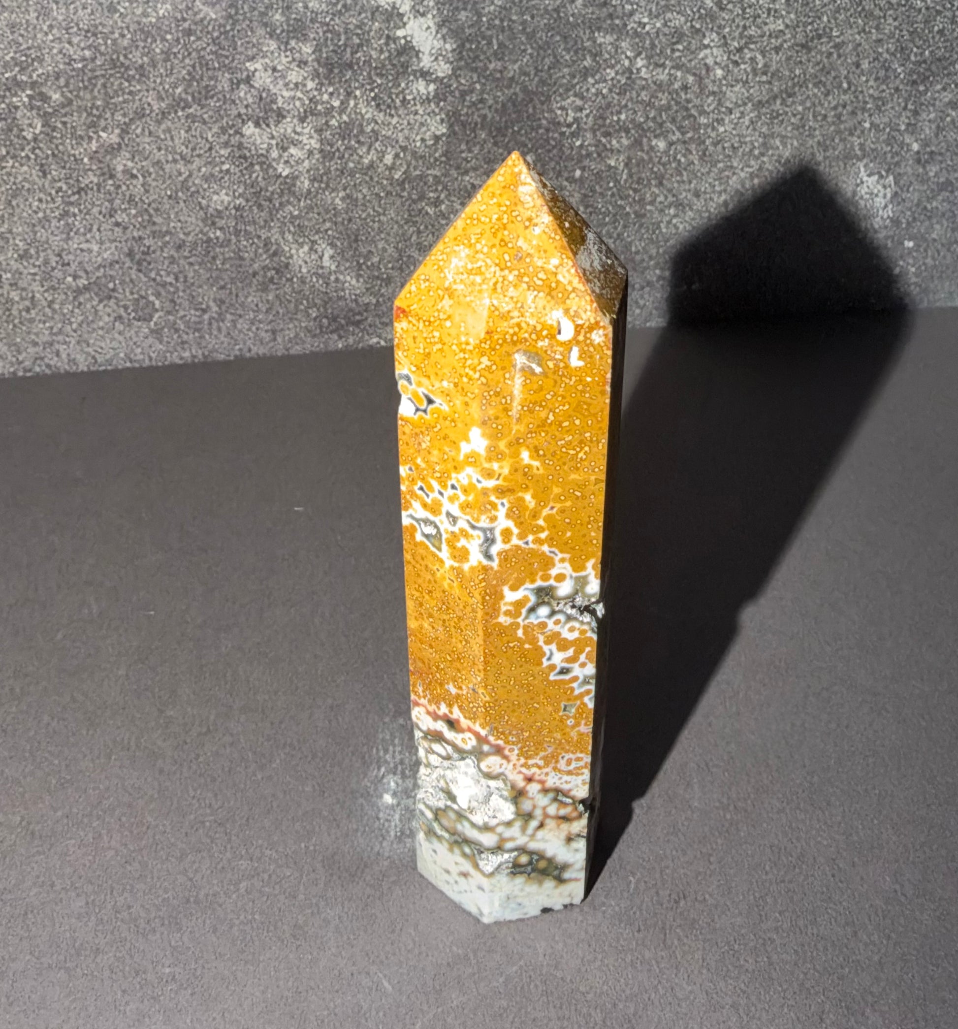 A polished golden-yellow crystal point with white and gray veining, standing upright on a dark gray surface against a textured gray background.