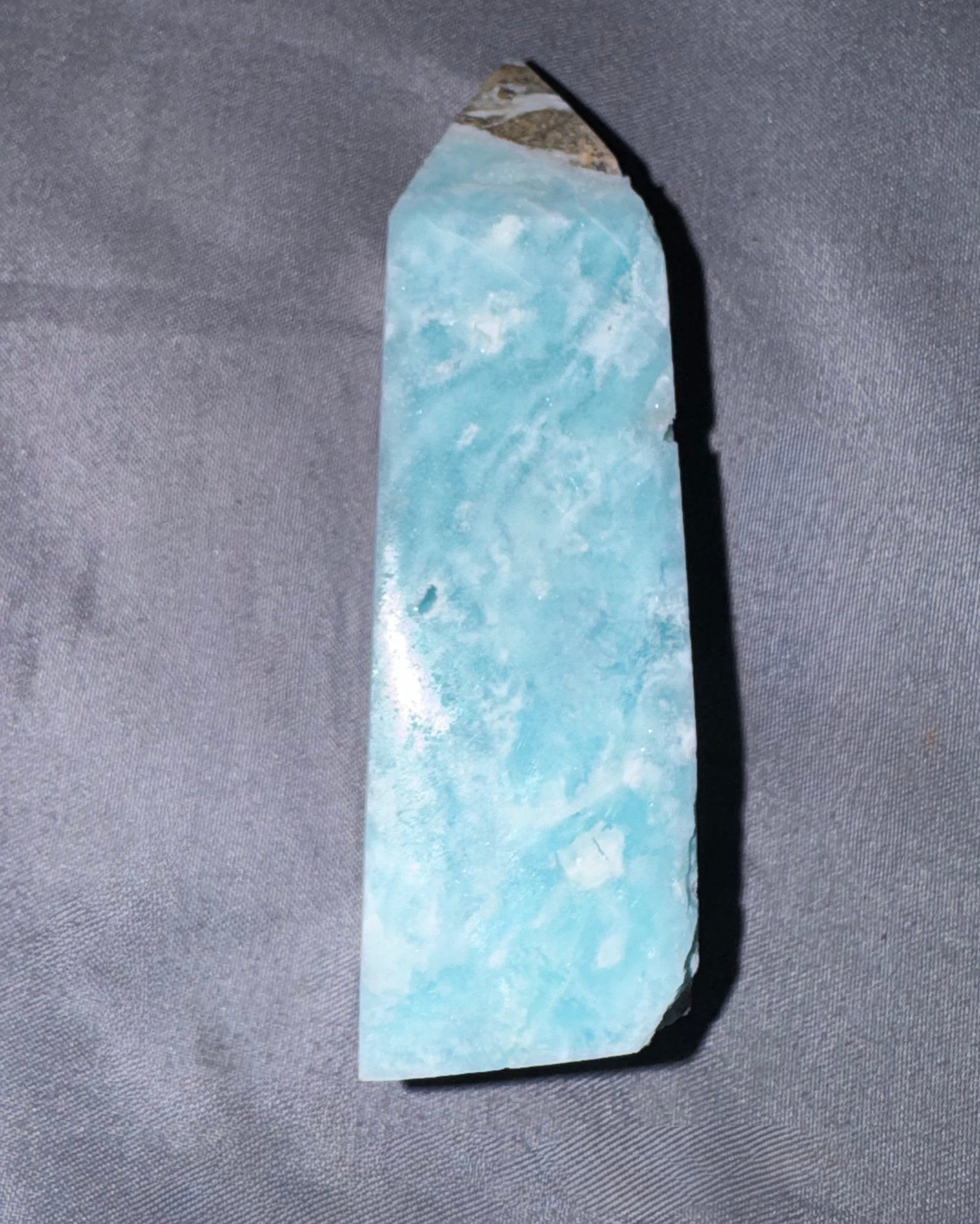A raw light blue  crystal point with a natural rough texture and a small brownish tip, standing vertically on a gray textured fabric background.