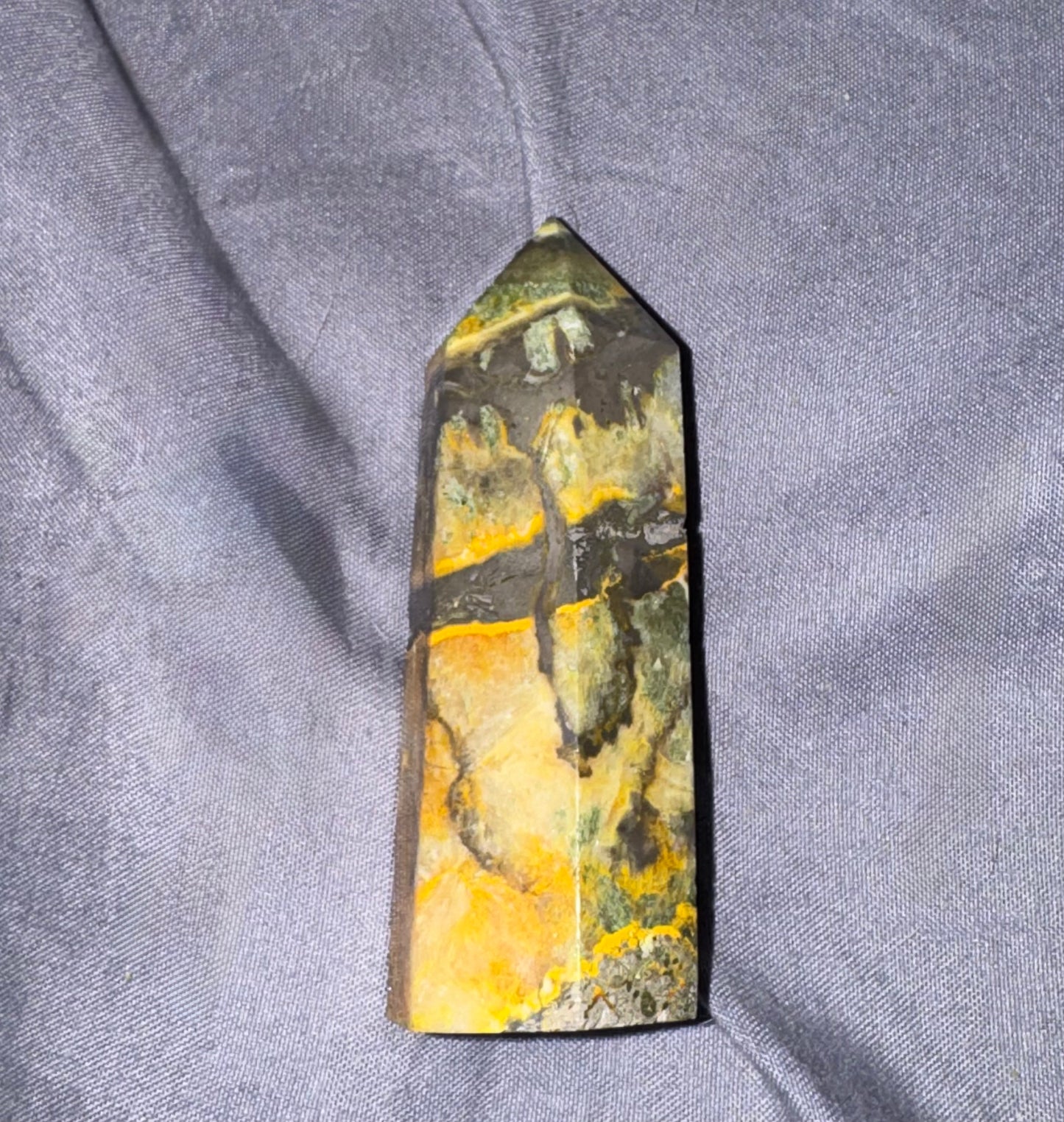 A polished pointed crystal tower, likely a jasper point, featuring natural marbled patterns of yellow, green, and dark gray, resting on a textured gray fabric background.