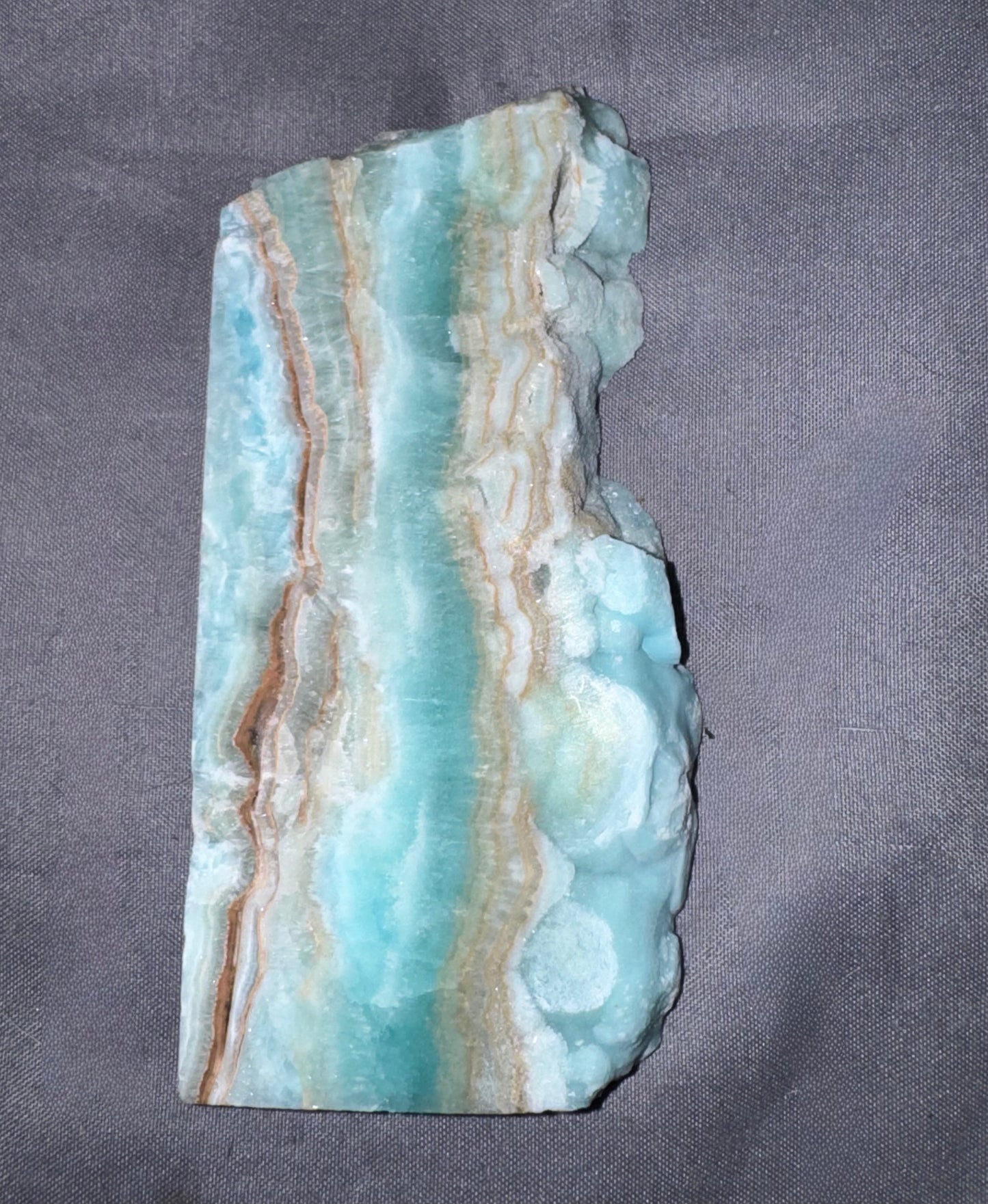 A polished natural stone slab with wavy bands of turquoise, light blue, beige, and brown, displayed on a textured gray fabric background.