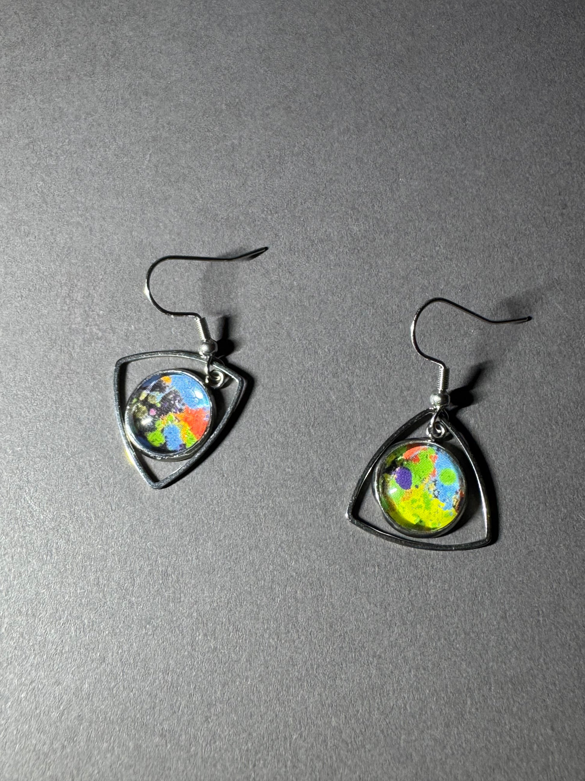 A pair of silver-toned dangle earrings with shield-shaped frames and oval colorful globe cabochons, displayed on a textured gray background.