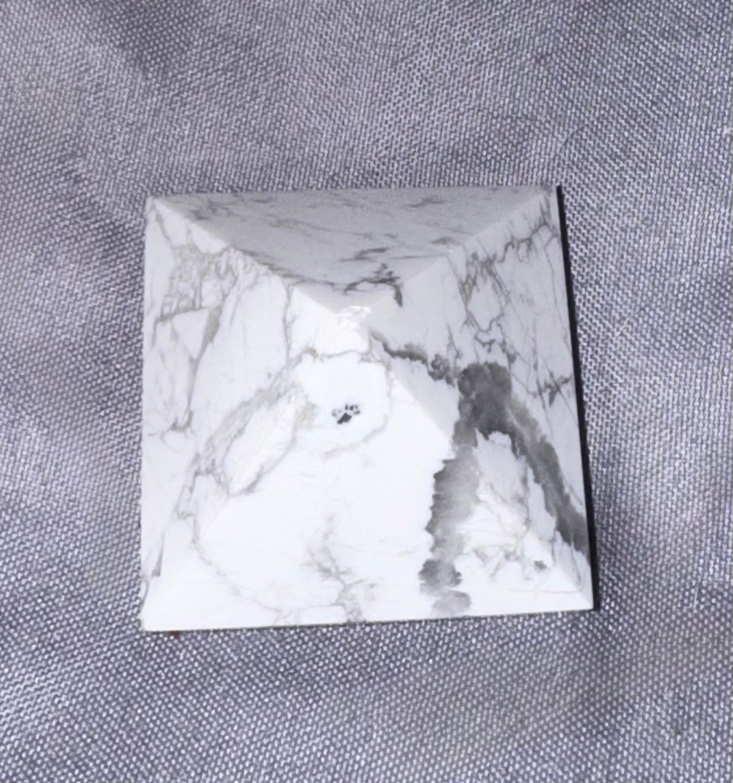 A square white marble or quartzite gemstone cabochon with gray veining, displayed on a textured gray fabric background.