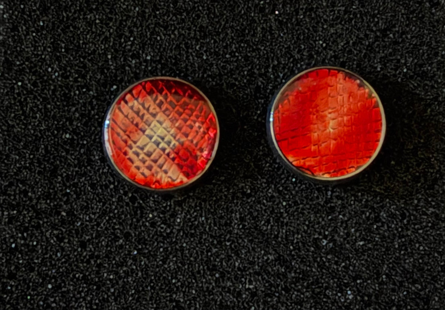 Two round red textured cabochon studs with silver metal borders, displayed side-by-side on a black textured background.