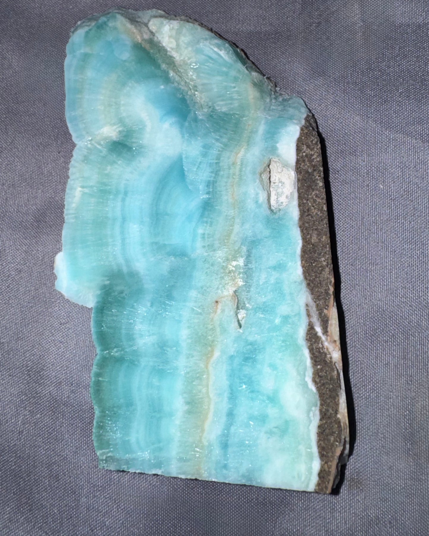 A raw mineral specimen of turquoise-blue barite with natural banding and a dark brown to black rough outer matrix, displayed on a textured gray fabric background.