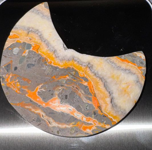 A crescent-shaped stone coaster featuring a marbled pattern of gray, orange, cream, and white, displayed on a black and brushed metal surface.