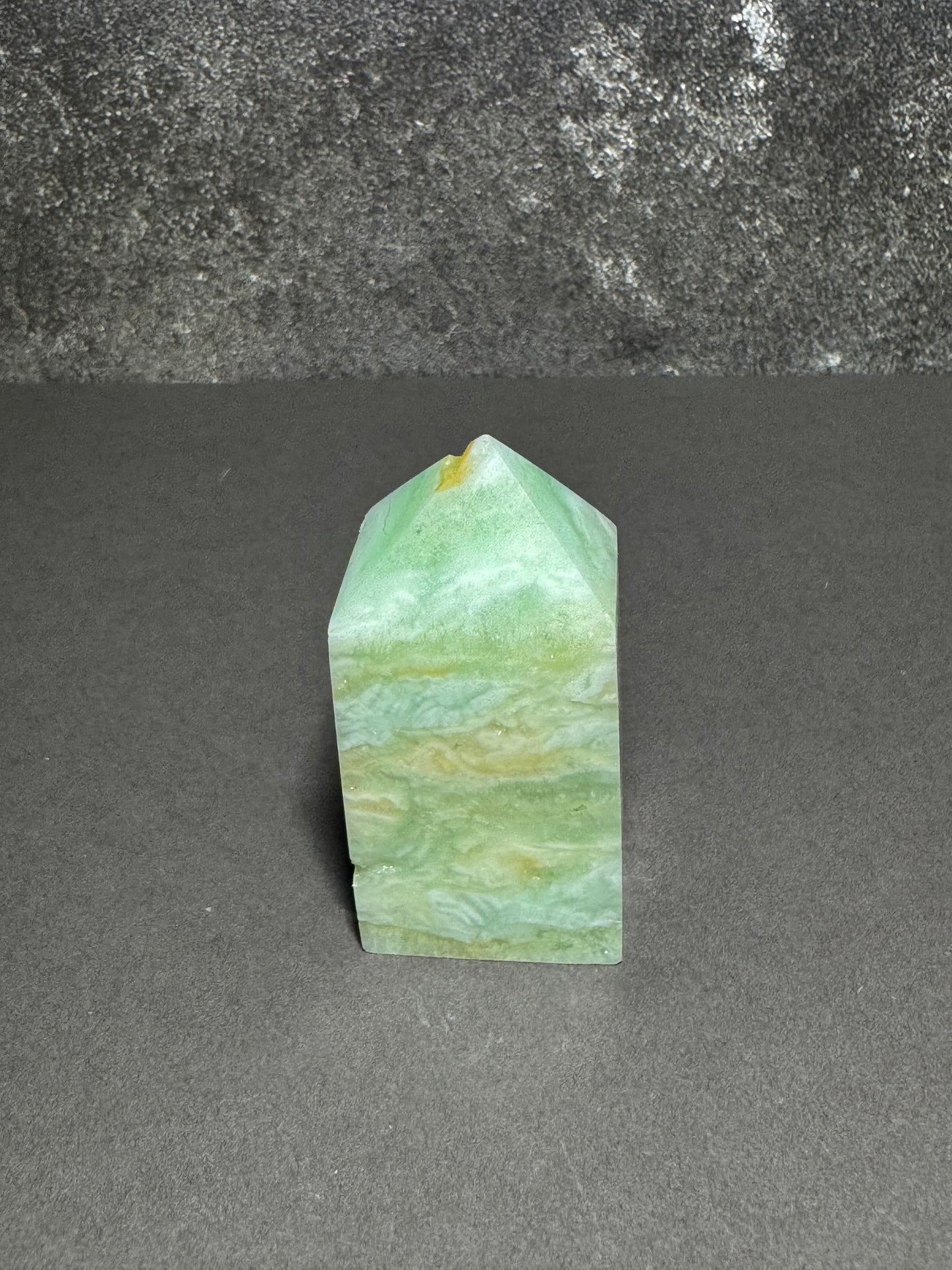 A natural green and white marbled crystal point tower, likely a jasper or quartzite point, standing upright on a dark gray surface against a textured gray background.