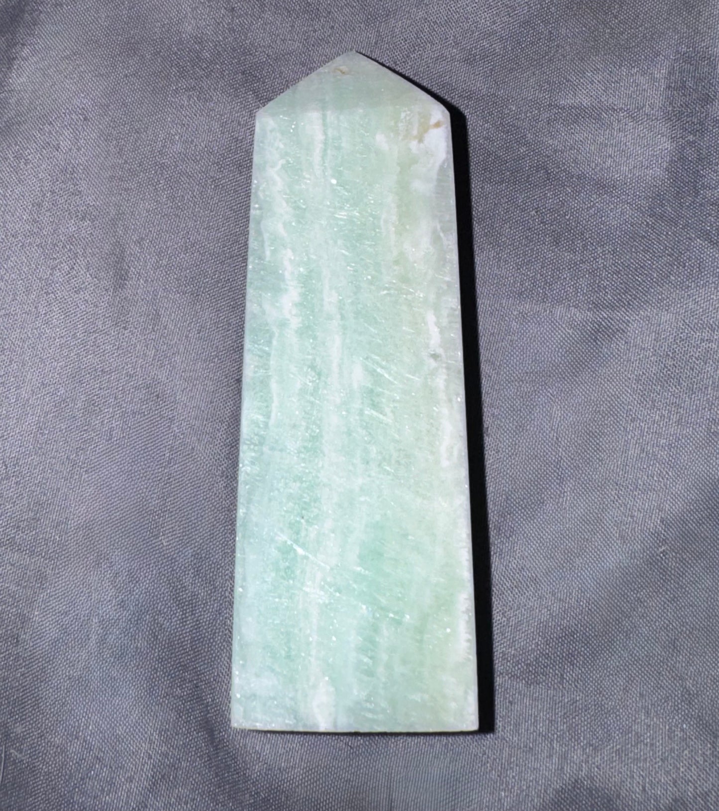 A pointed rectangular stone or crystal with a pale green and white marbled texture, photographed upright on a textured gray fabric background.
