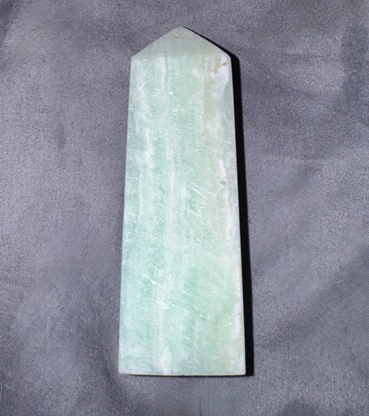A pointed rectangular stone or crystal with a pale green and white marbled texture, photographed upright on a textured gray fabric background.