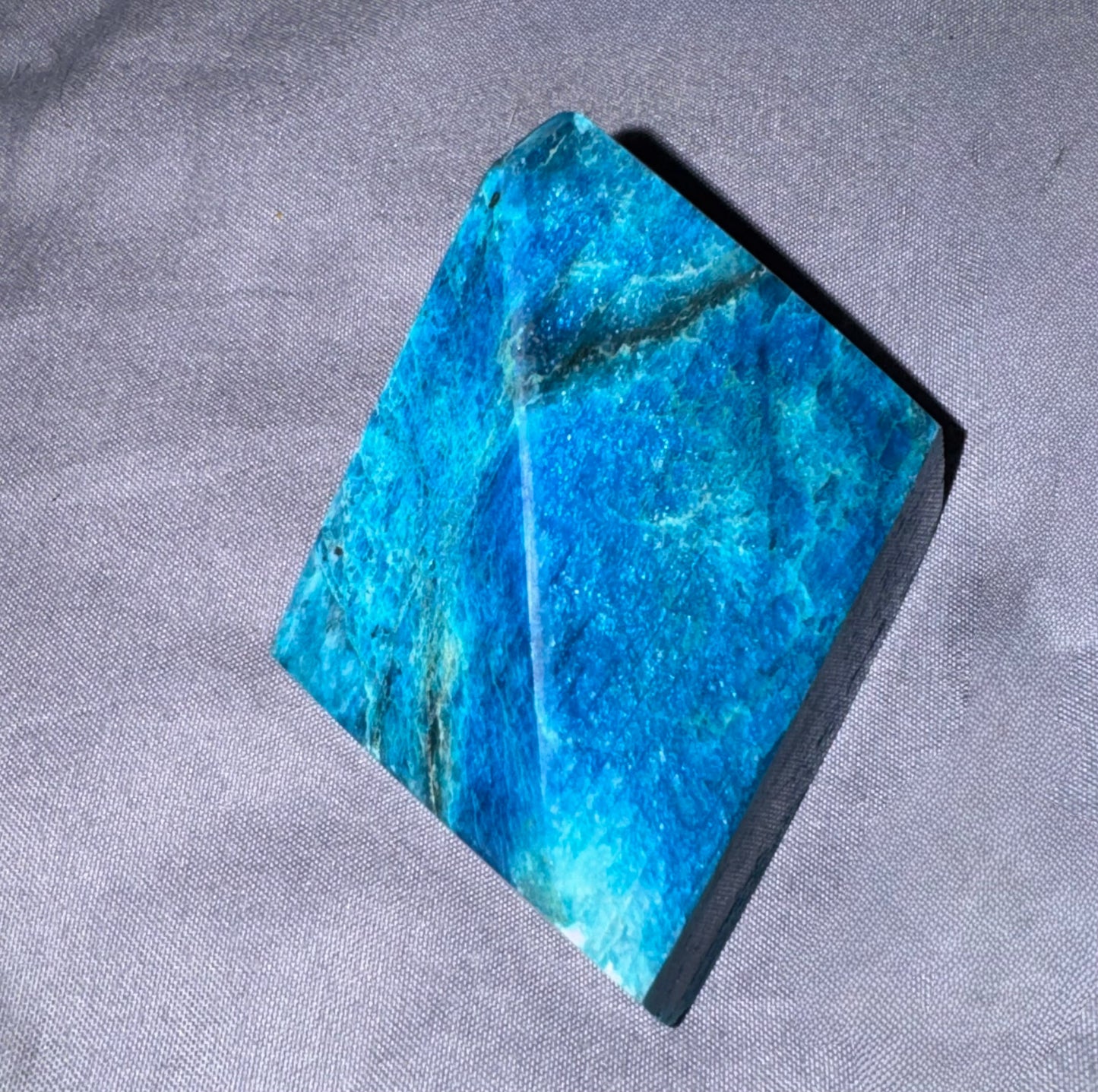 A polished blue stone or crystal with vivid turquoise and teal tones, photographed on a textured gray fabric background.