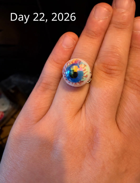 A close-up of a hand wearing a unique ring shaped like a realistic human eye, with the text 'Day 22, 2026' overlaid at the top left.
