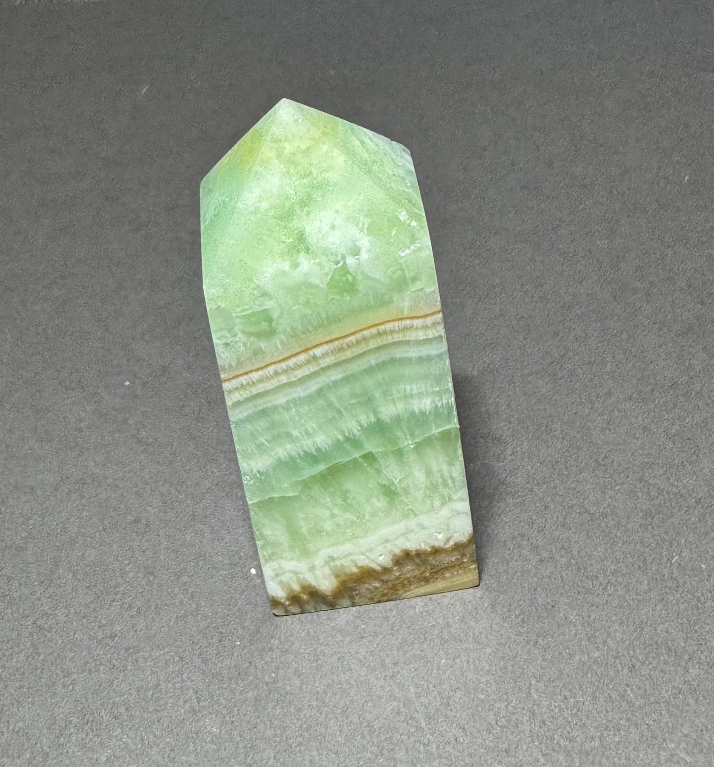 A natural polished crystal point featuring light green, white, and brown banding, standing upright on a neutral gray surface.