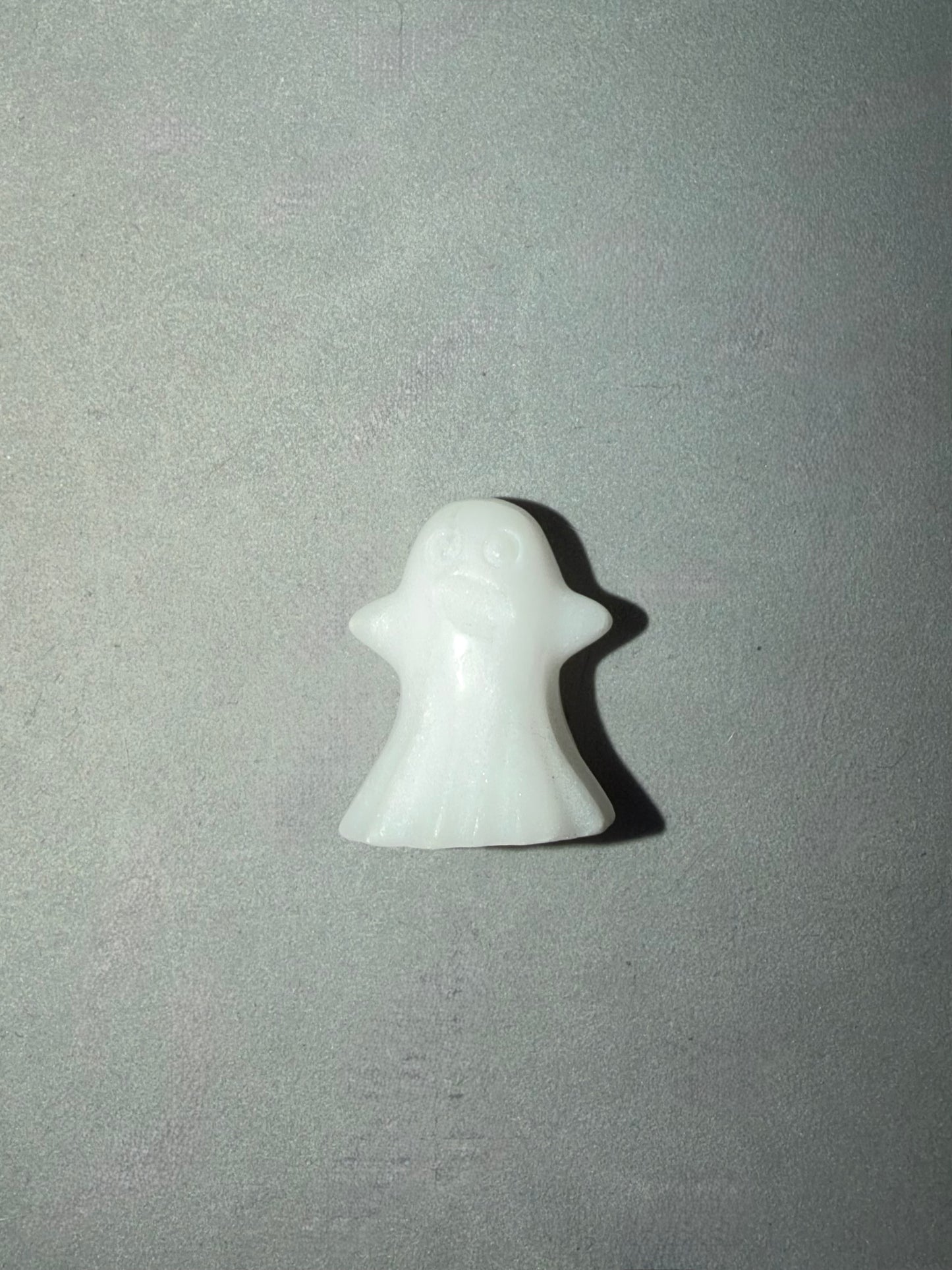 A small white ghost-shaped figurine or charm with a smooth glossy finish, centered on a textured gray background.