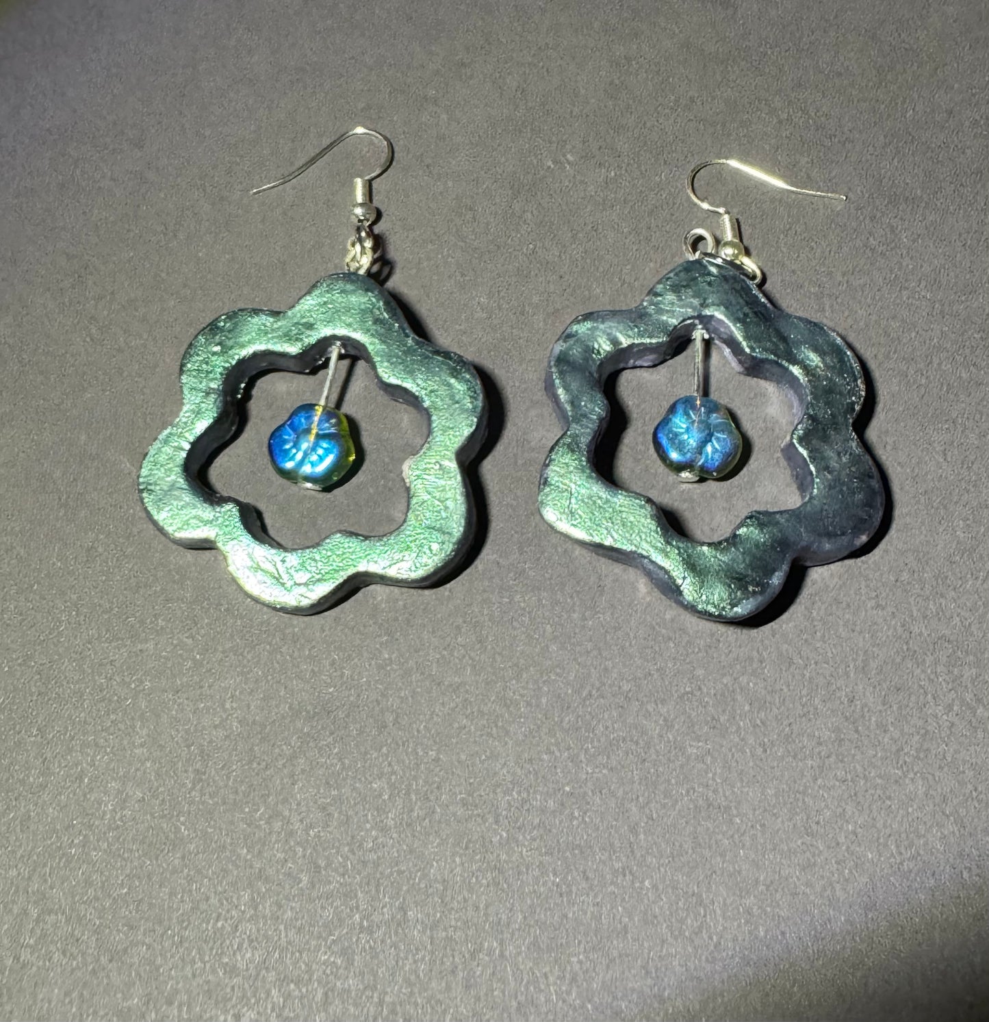 A pair of handmade dangle earrings with textured, scalloped silver-tone metal frames and small blue glass beads inside, shown on a neutral gray background.