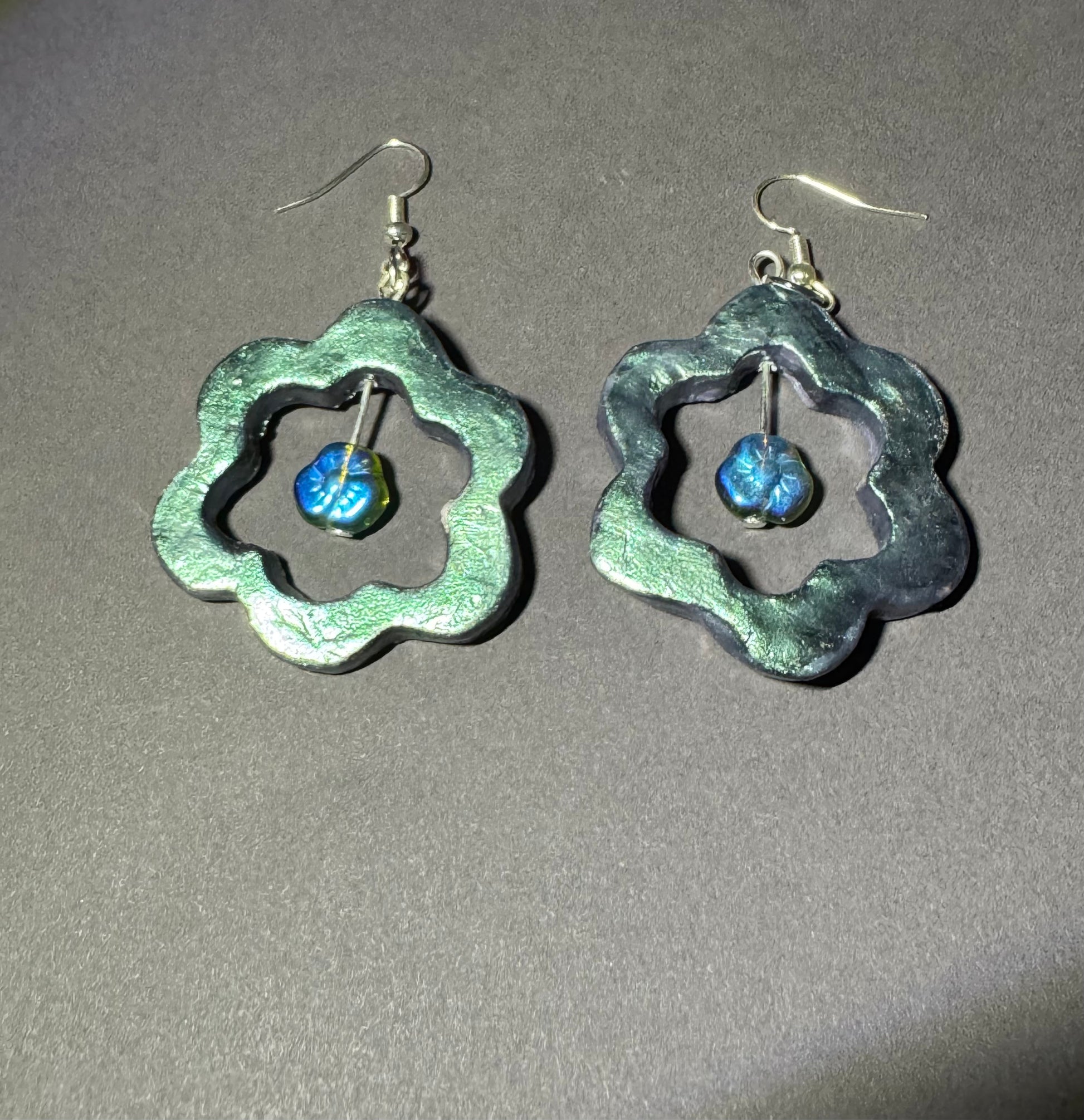 A pair of handmade dangle earrings with textured, scalloped silver-tone metal frames and small blue glass beads inside, shown on a neutral gray background.