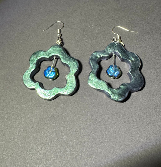 A pair of handmade dangle earrings with textured, scalloped silver-tone metal frames and small blue glass beads inside, shown on a neutral gray background.