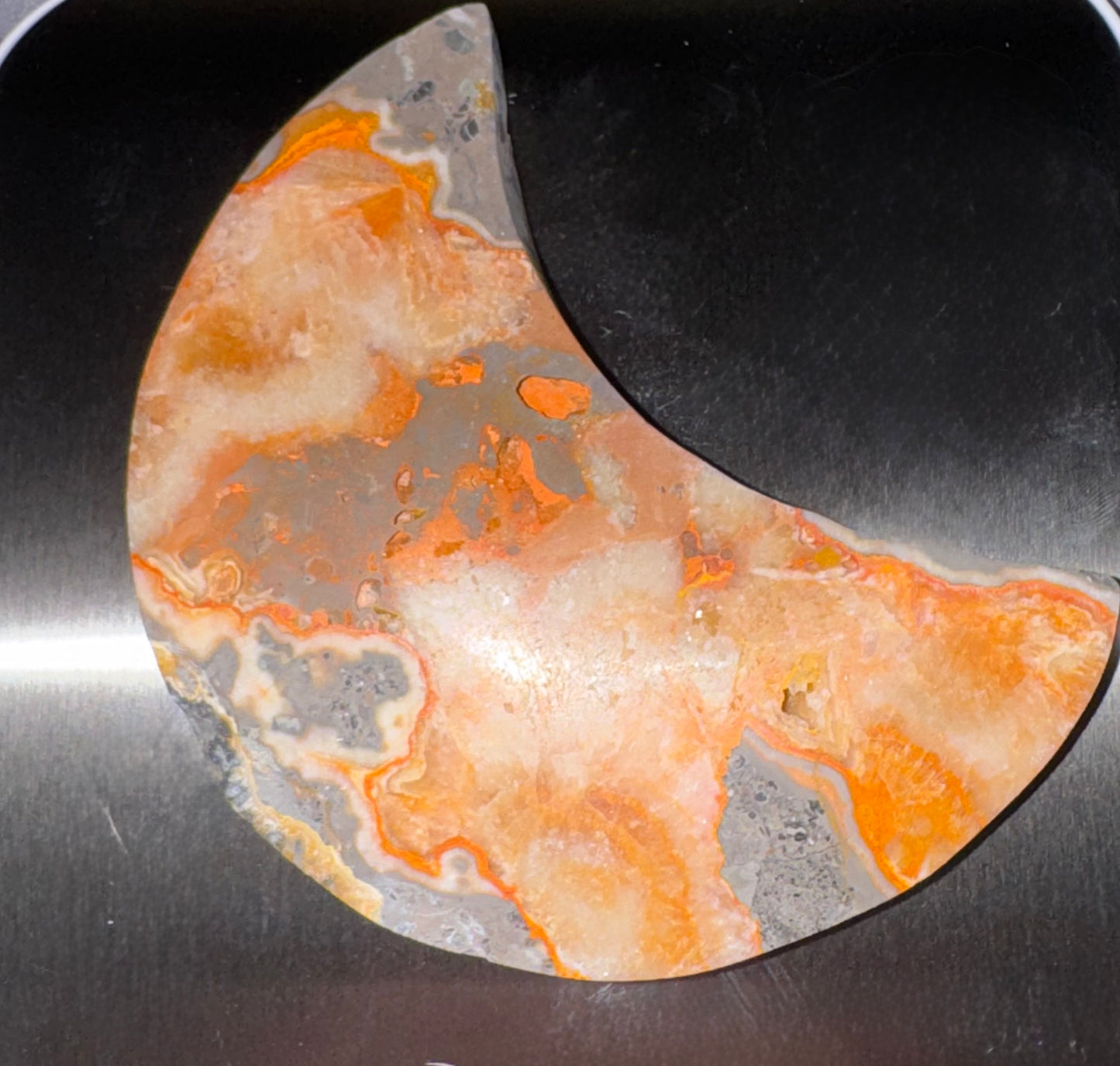 A crescent-shaped decorative stone or acrylic piece with marbled orange, cream, and gray patterns, shown on a dark metallic surface.