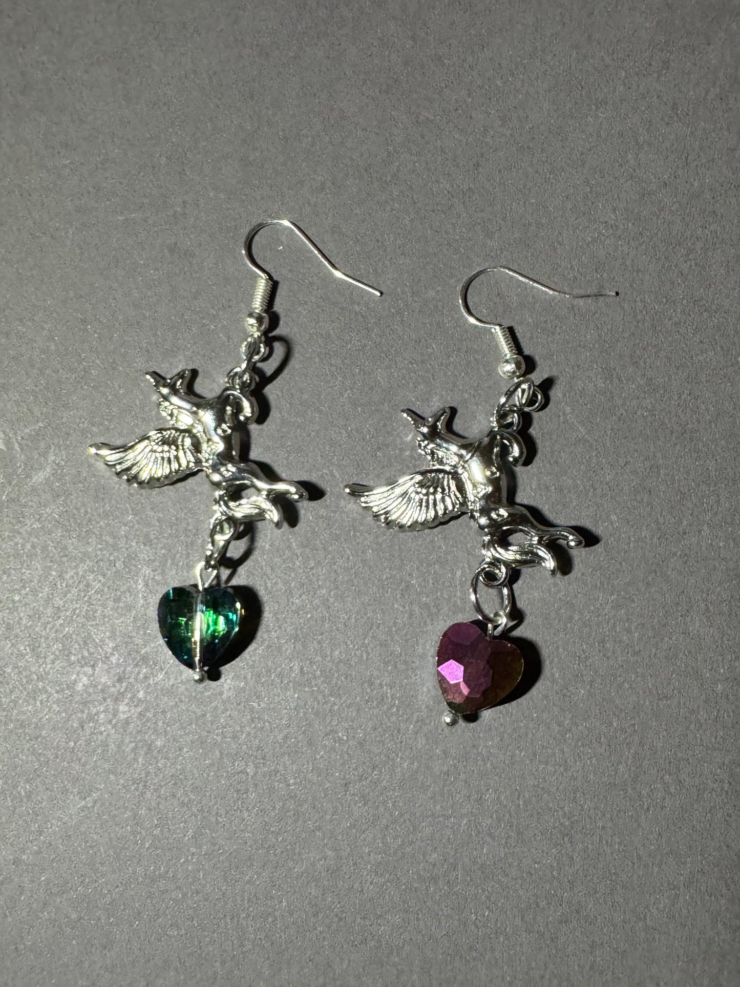 A pair of silver-tone dangling earrings featuring winged cherub charms and heart-shaped dangling stones, one green and one pink, shown on a plain gray background.