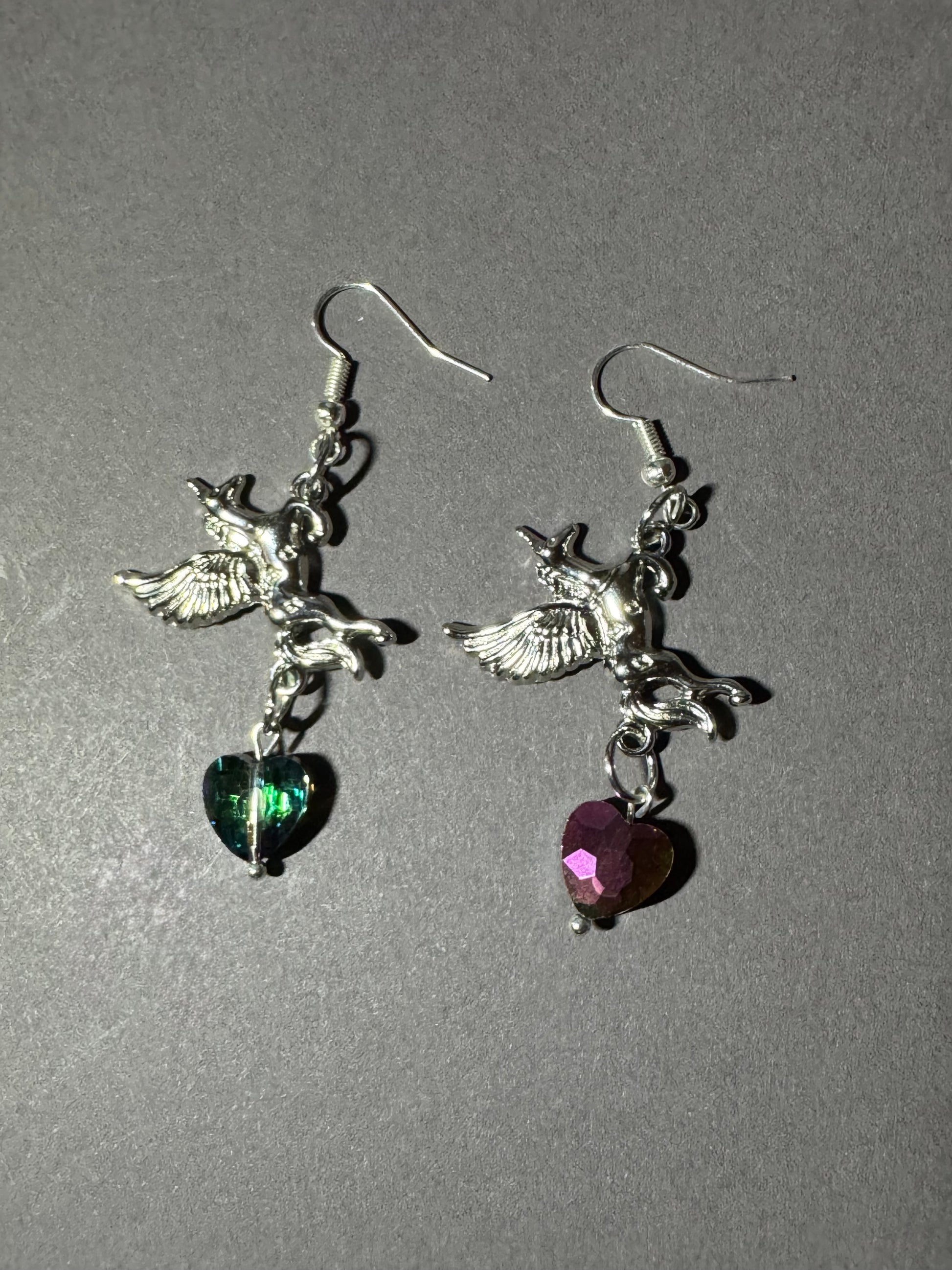 A pair of silver-tone dangling earrings featuring winged cherub charms and heart-shaped dangling stones, one green and one pink, shown on a plain gray background.