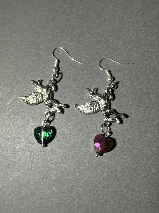 A pair of silver-tone dangling earrings featuring winged cherub charms and heart-shaped dangling stones, one green and one pink, shown on a plain gray background.