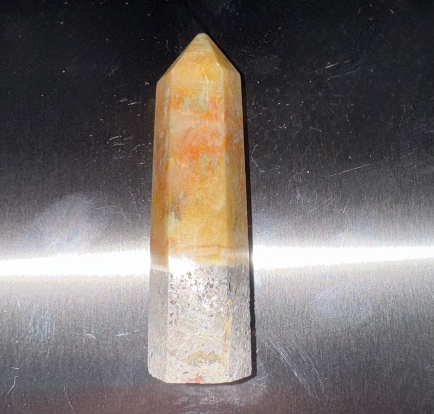 A natural quartz crystal point with a dual-layered design, featuring a warm orange and yellow top section and a clear, textured white bottom section, set against a reflective metallic background.