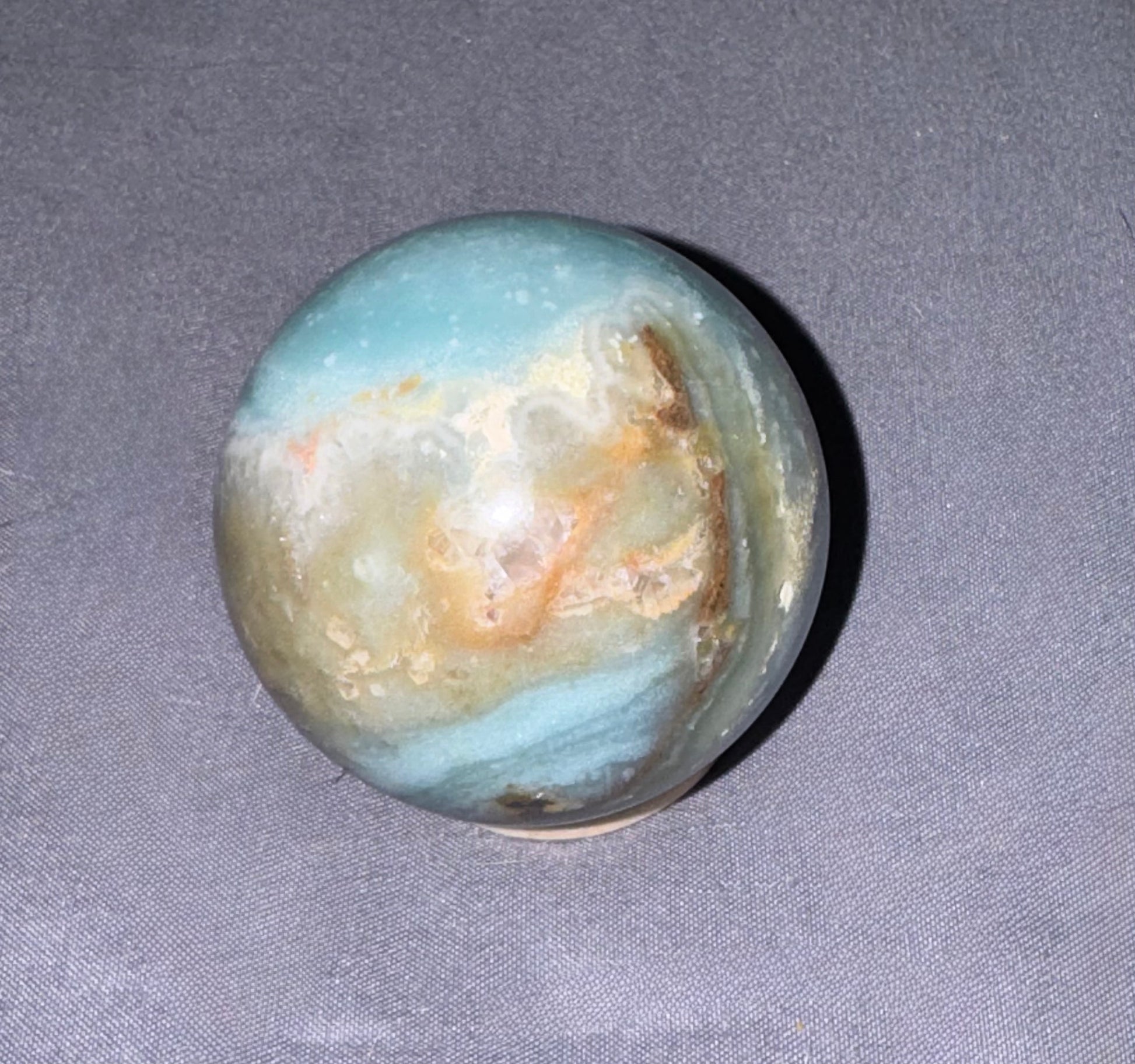 A polished round crystal sphere with natural banding of light blue, cream, tan, and brown, resting on a textured gray fabric background.
