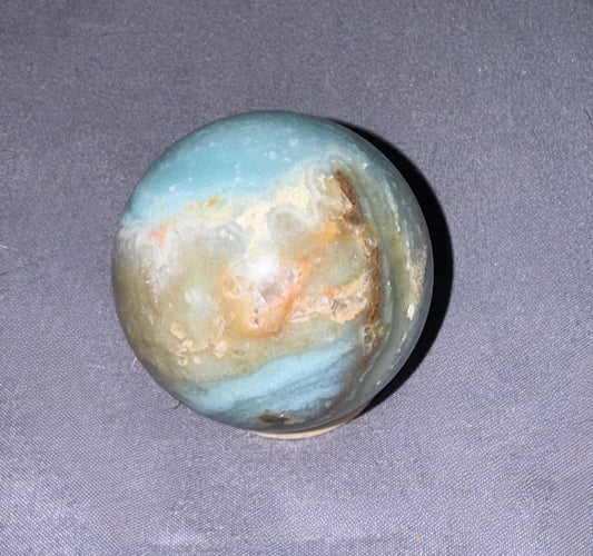 A polished round crystal sphere with natural banding of light blue, cream, tan, and brown, resting on a textured gray fabric background.