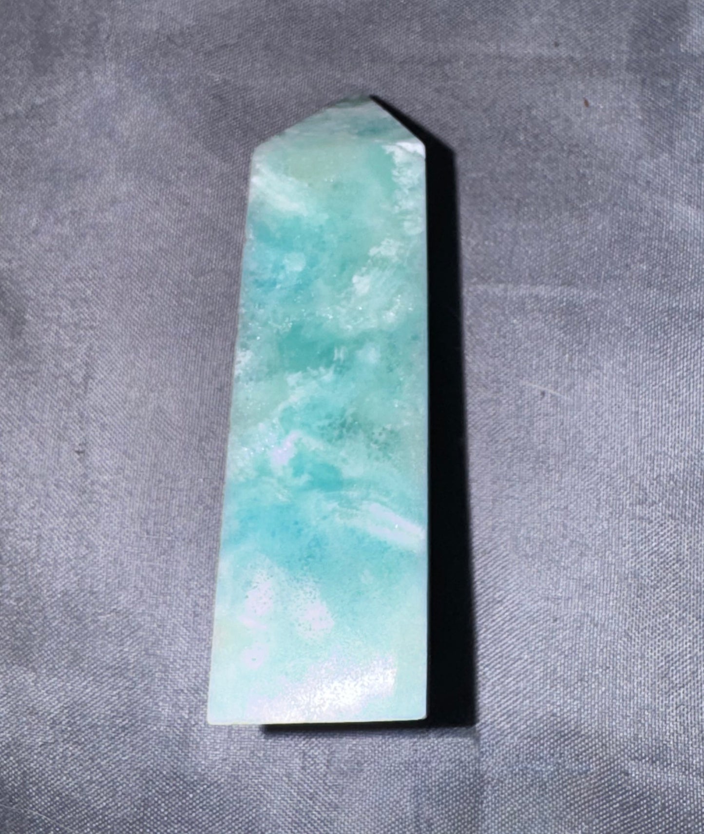 A polished Amazonite crystal point with natural light green and turquoise marbling, resting on a textured gray fabric background.