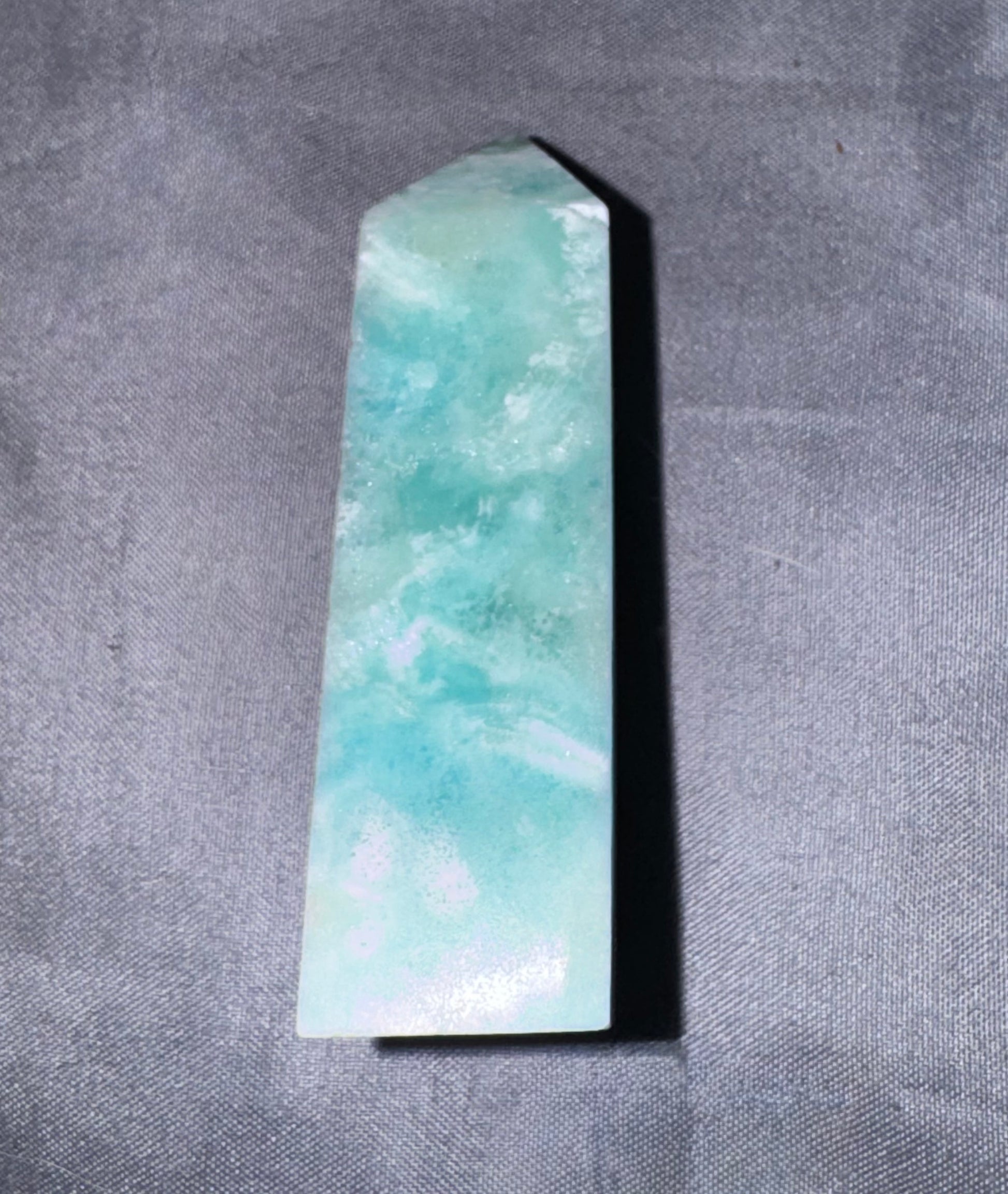 A polished Amazonite crystal point with natural light green and turquoise marbling, resting on a textured gray fabric background.